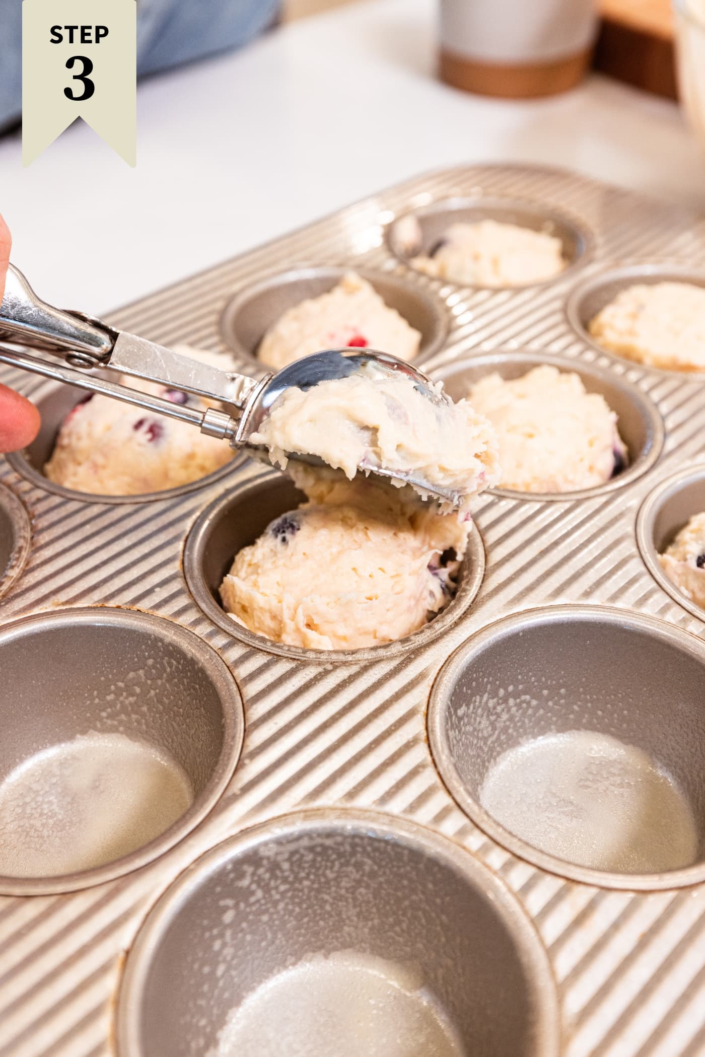 Muffin tins filled with blueberry muffin batter with scoop scooping batter into tins.