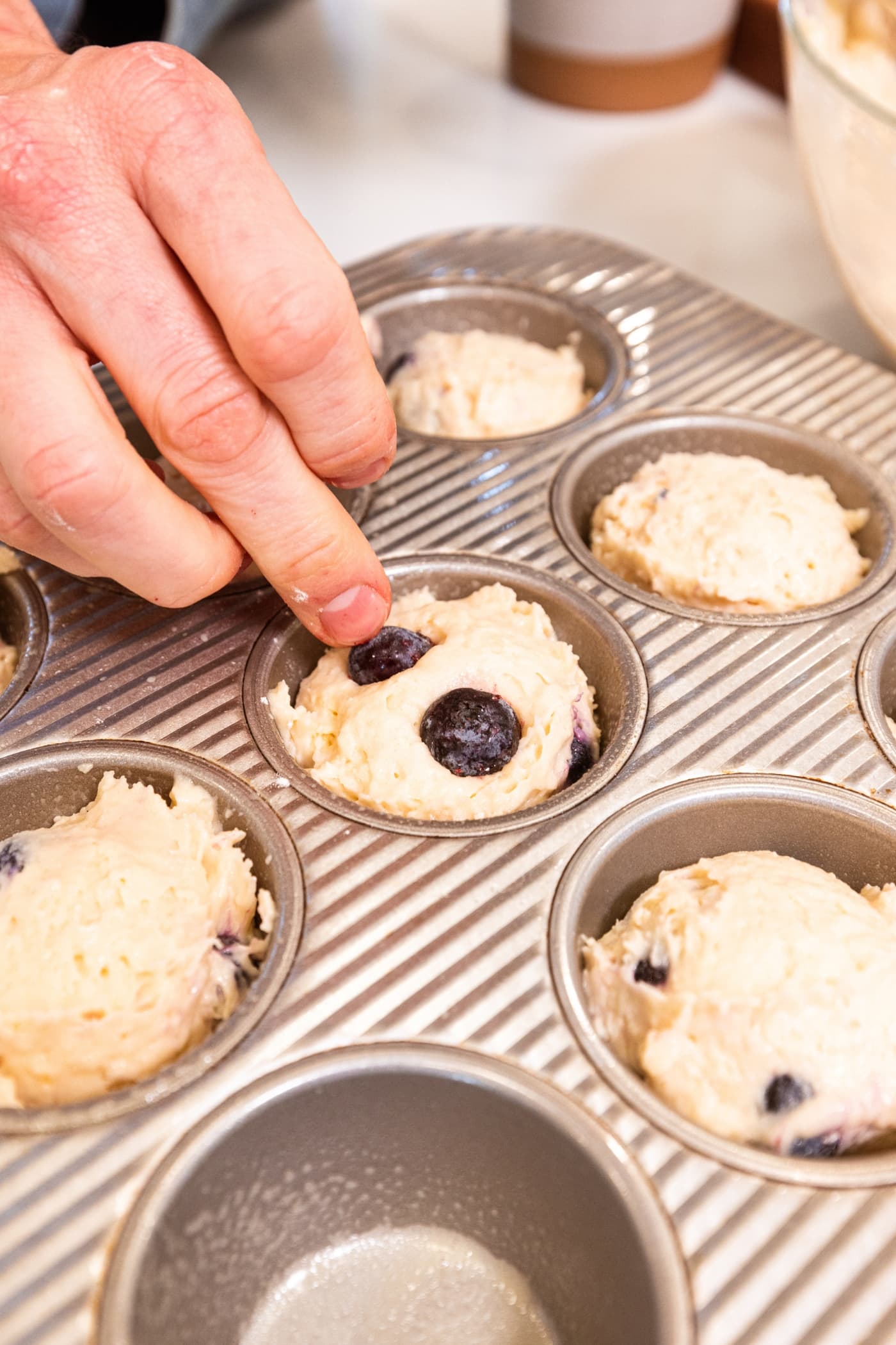 Hand placing blueberries on top of batter in muffin tins for blueberry muffins.