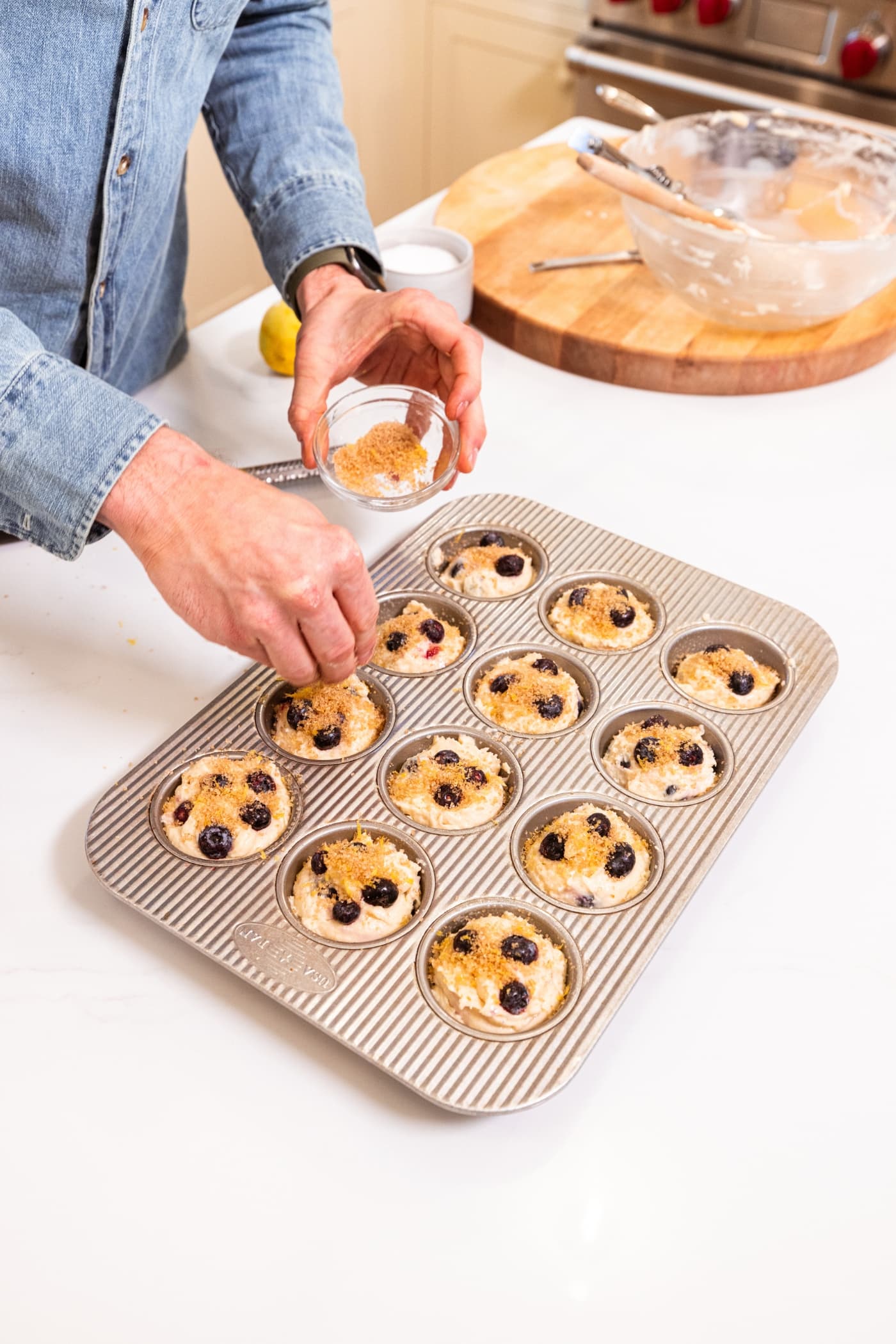 Hand holding small bowl filled with mixture of lemon zest and turbinado sugar sprinkling on top of blueberry muffins before being baked.