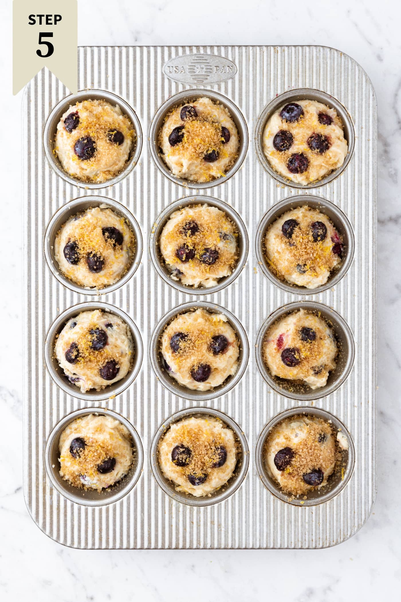 Metal muffin tin sitting on marble surface filled with unbaked batter for blueberry muffins.