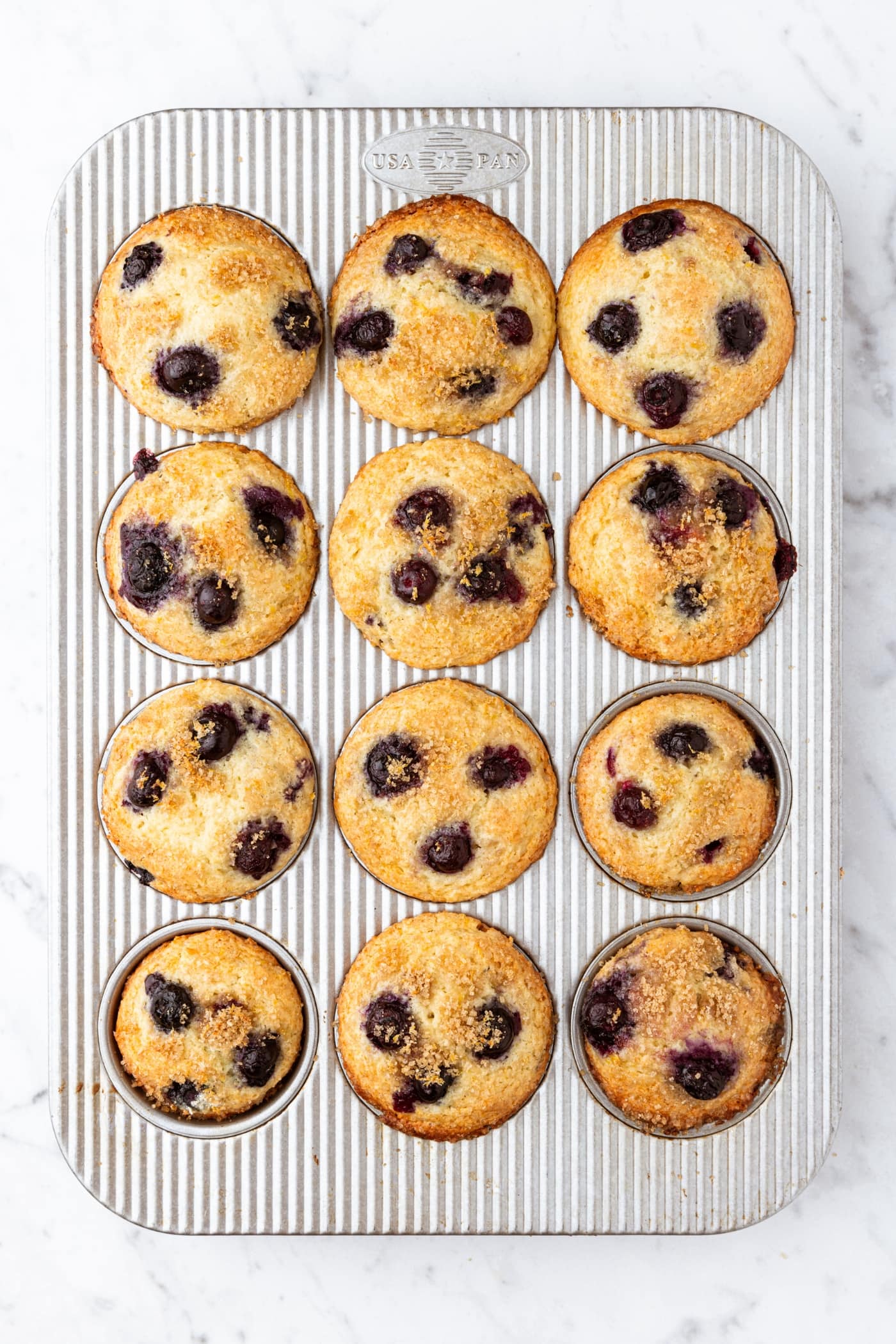 White marble surface with metal muffin tin with freshly baked blueberry muffins with lemon zest topping.