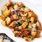 White oval serving platter filled with crisped rosemary roasted potatoes topped with herbs and shredded cheese all on white marble surface.
