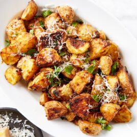 White oval serving platter filled with crisped rosemary roasted potatoes topped with herbs and shredded cheese all on white marble surface.