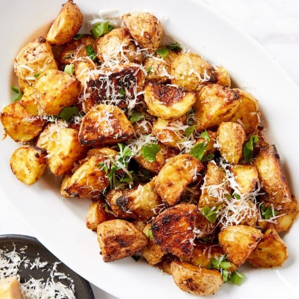 White oval serving platter filled with crisped rosemary roasted potatoes topped with herbs and shredded cheese all on white marble surface.