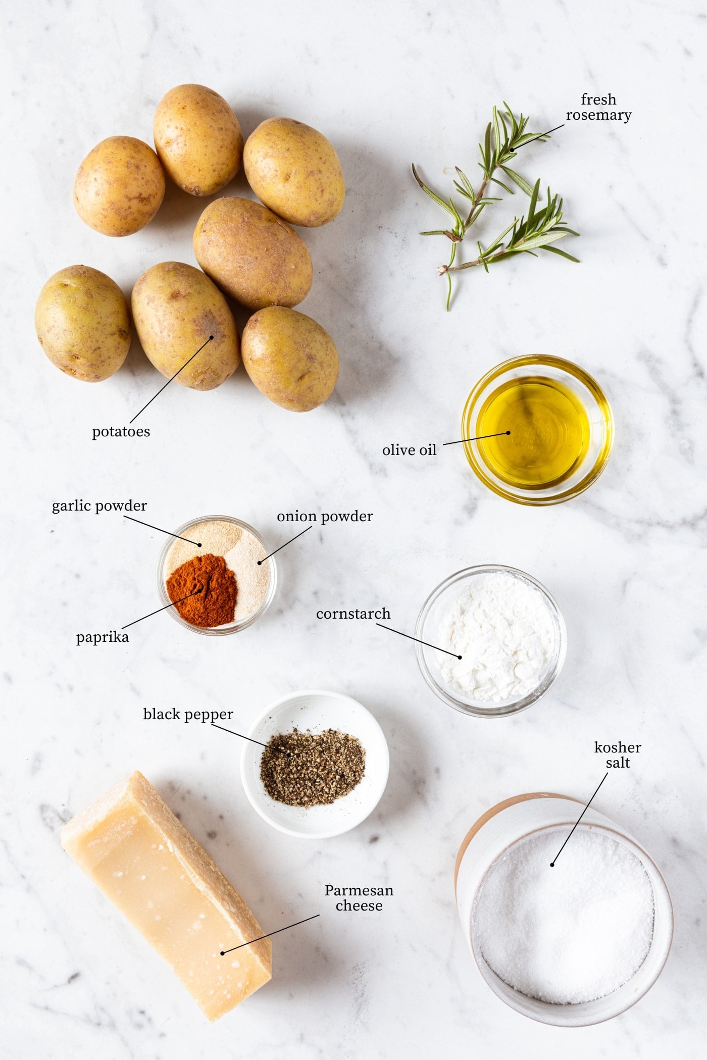 White marble surface filled with ingredients needed for rosemary roasted potatoes including small potatoes, spices, oil, cheese, and herbs.