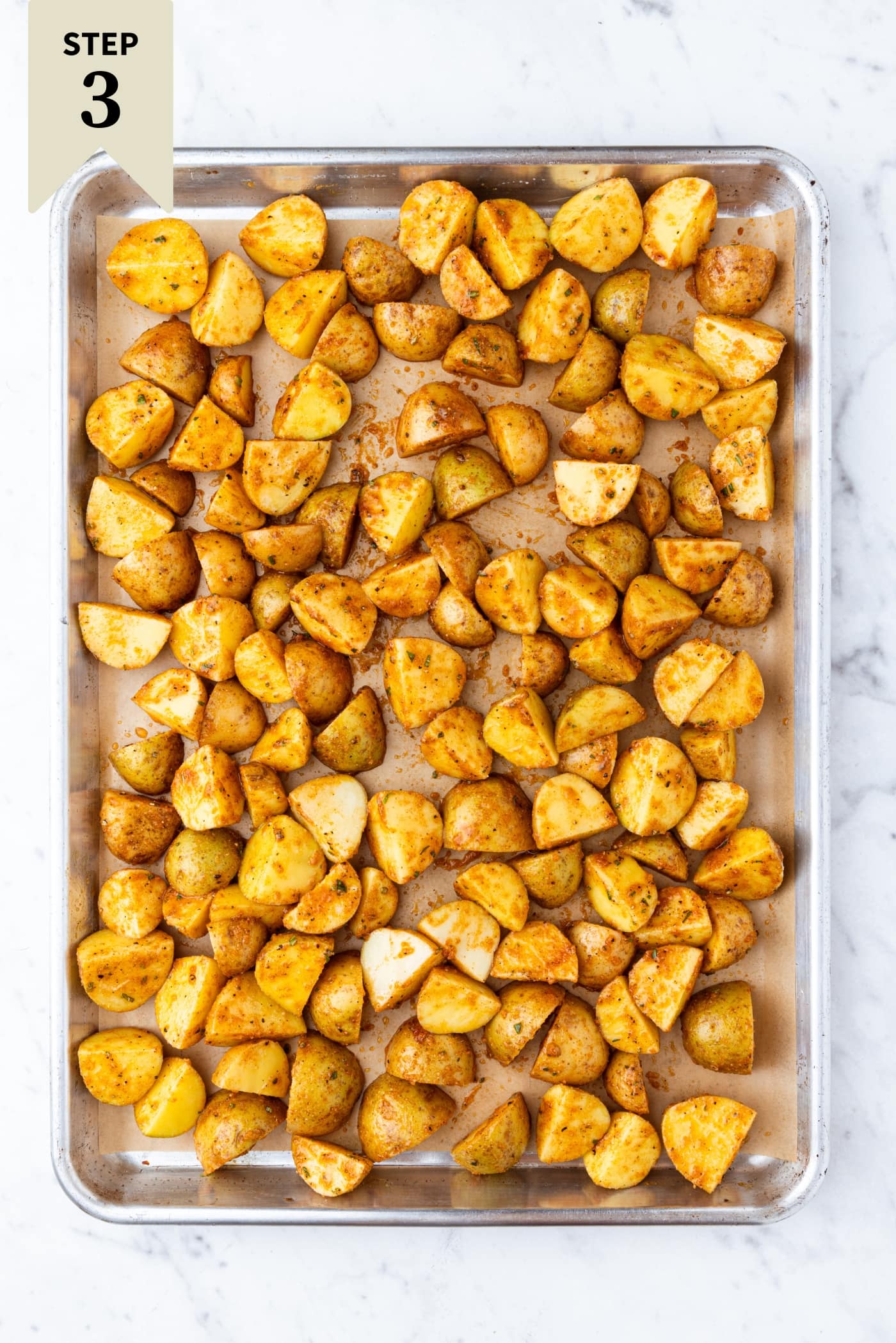 White marble surface with baking sheet with unbaked sliced potatoes with spices all over the top right before being baked.