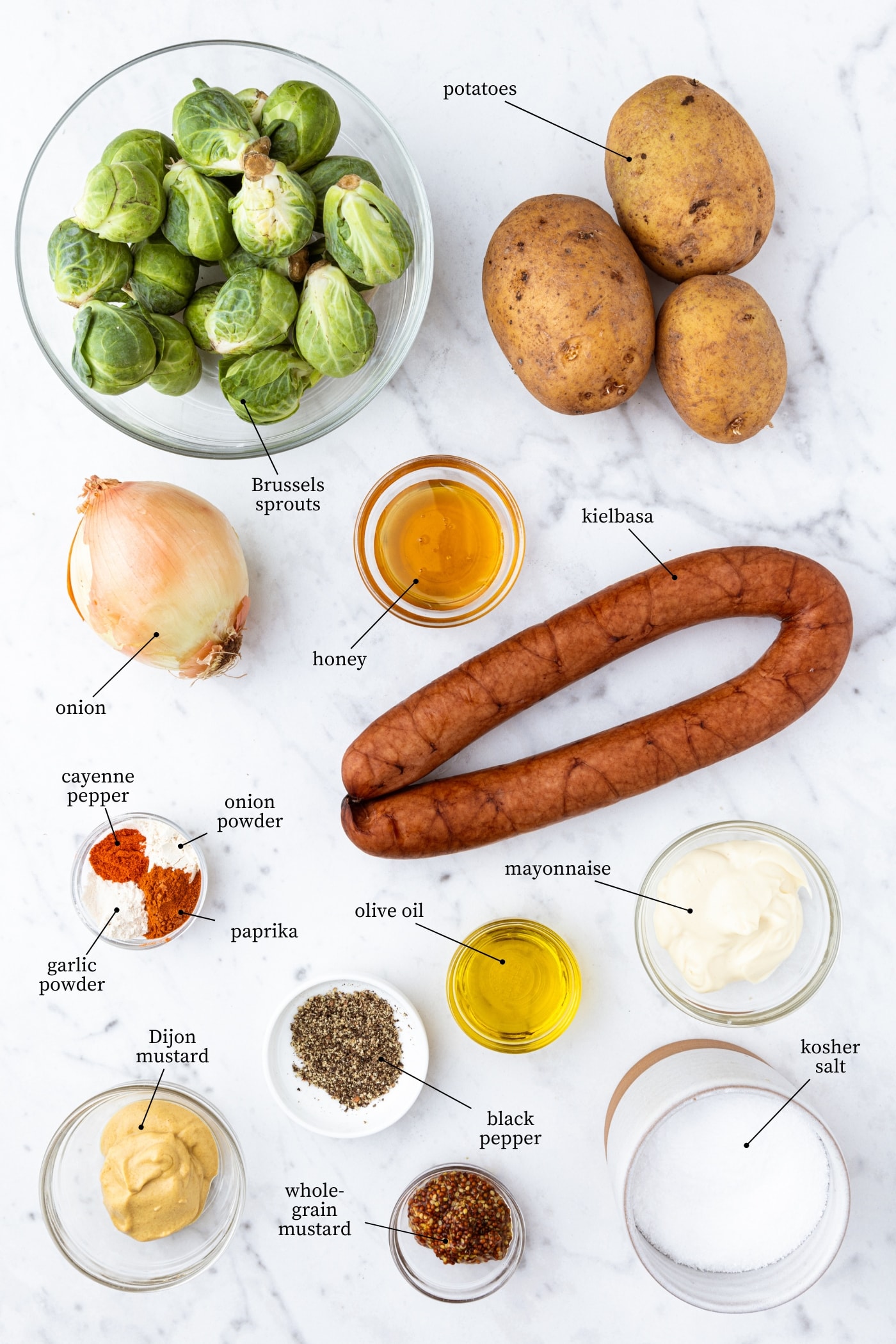 White marble surface filled with ingredients needed to make a kielbasa skillet meal including Brussels sprouts, potatoes, kielbasa, spices, and more.
