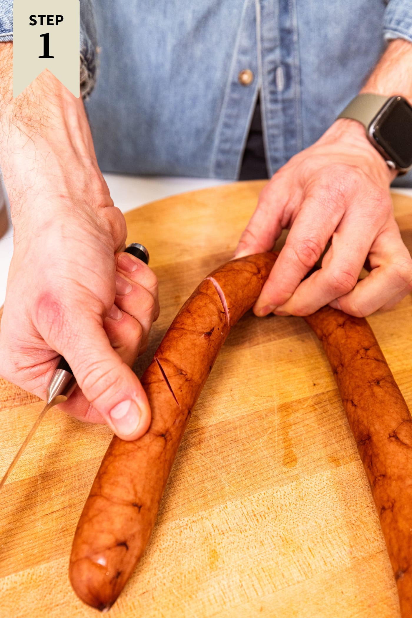Two hands holding kielbasa showing cuts that were made to score the piece of meat on wooden cutting board.