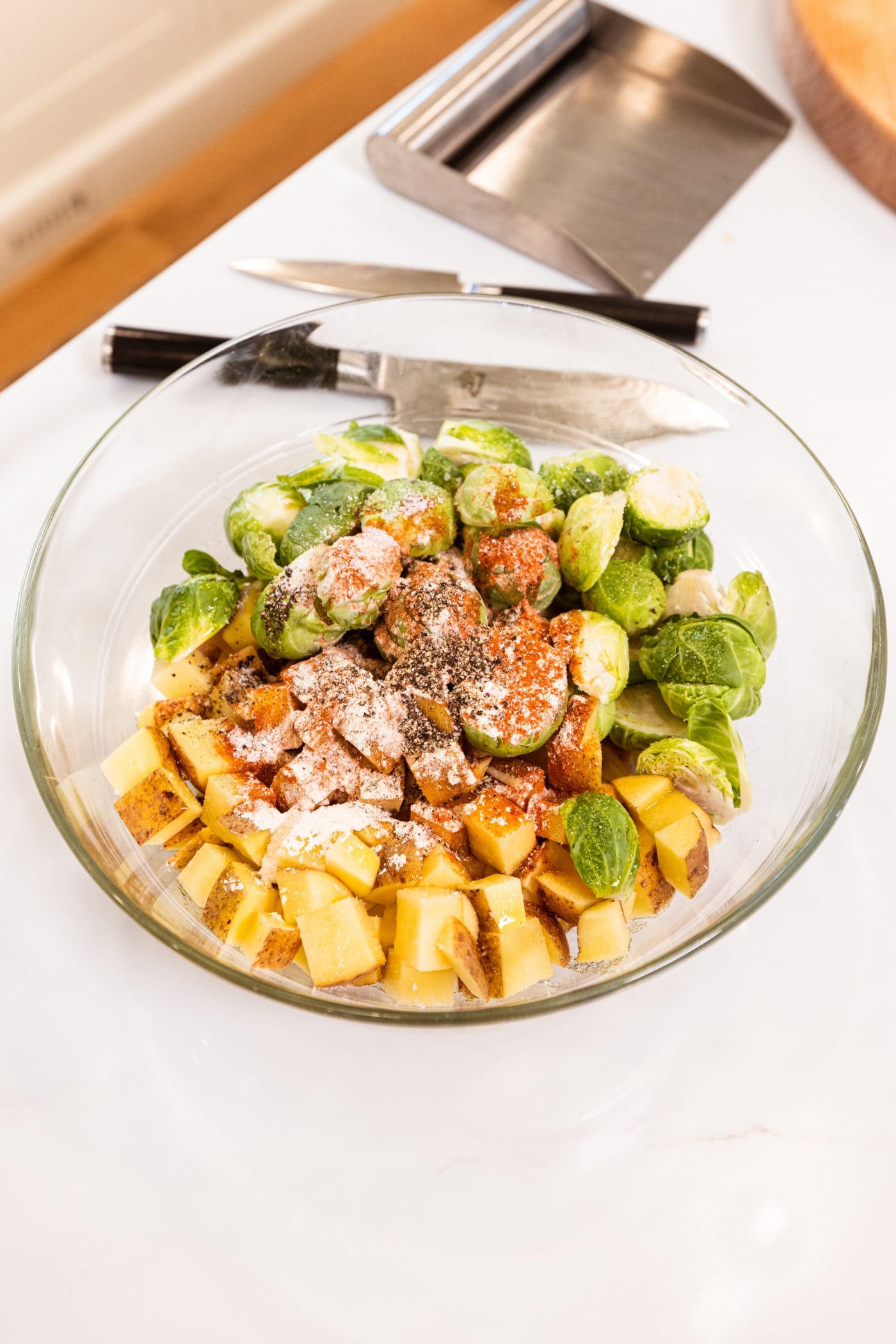 Glass bowl on white surface filled with sliced Brussels sprouts and potatoes with spices all over before being mixed together.