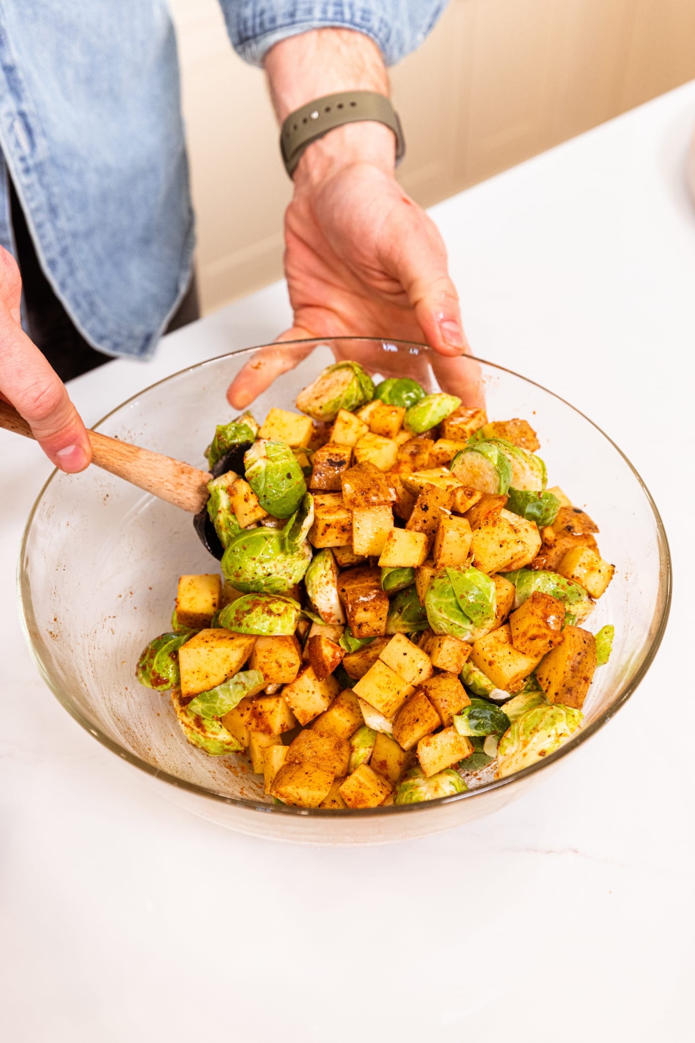 Hand using a spatula to mix together spices with sliced Brussels sprouts and potatoes in a glass bowl on white surface.