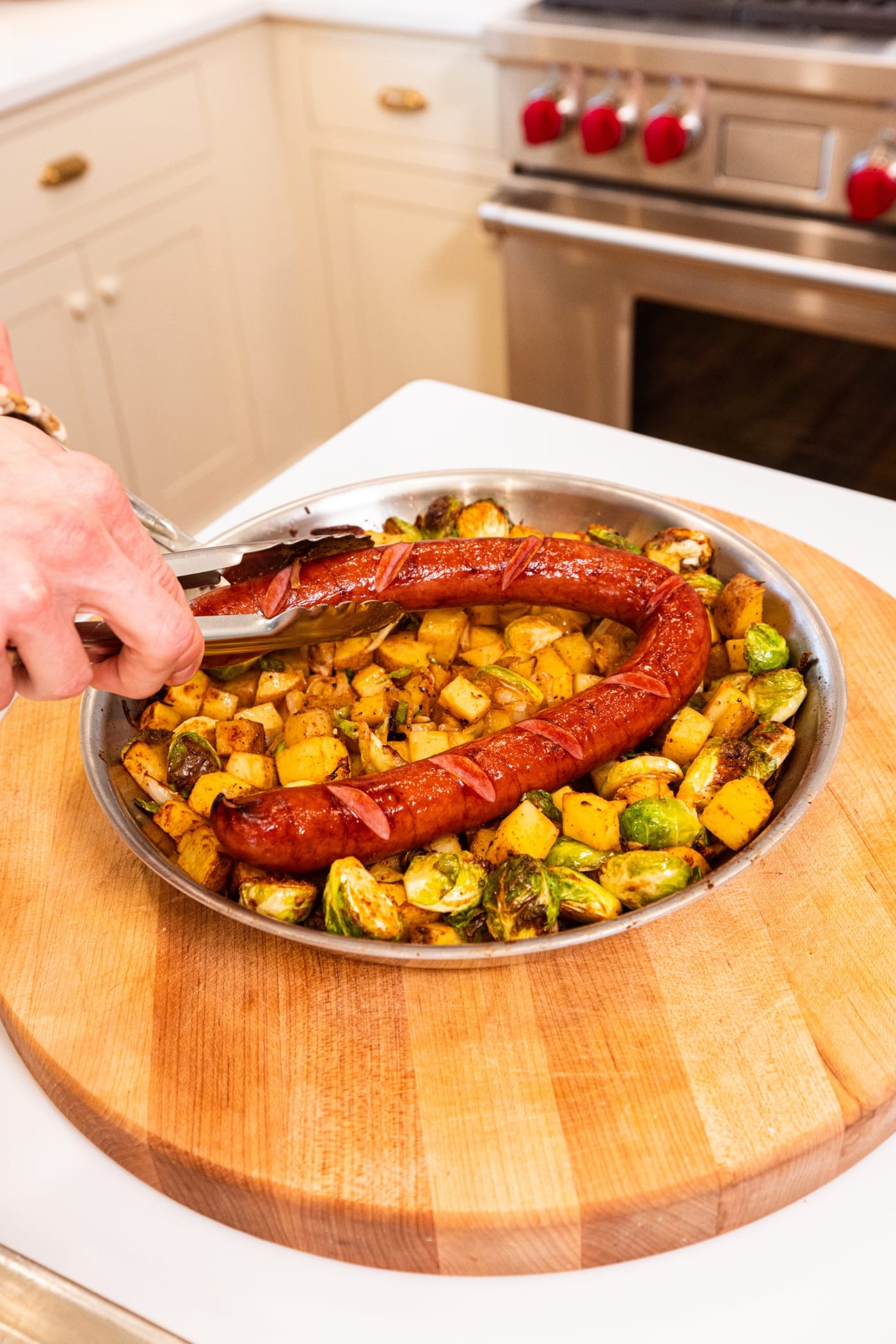 Hand using tongs to place a browned piece of kielbasa in a metal skillet with sliced Brussels sprouts and potatoes with spices all over.