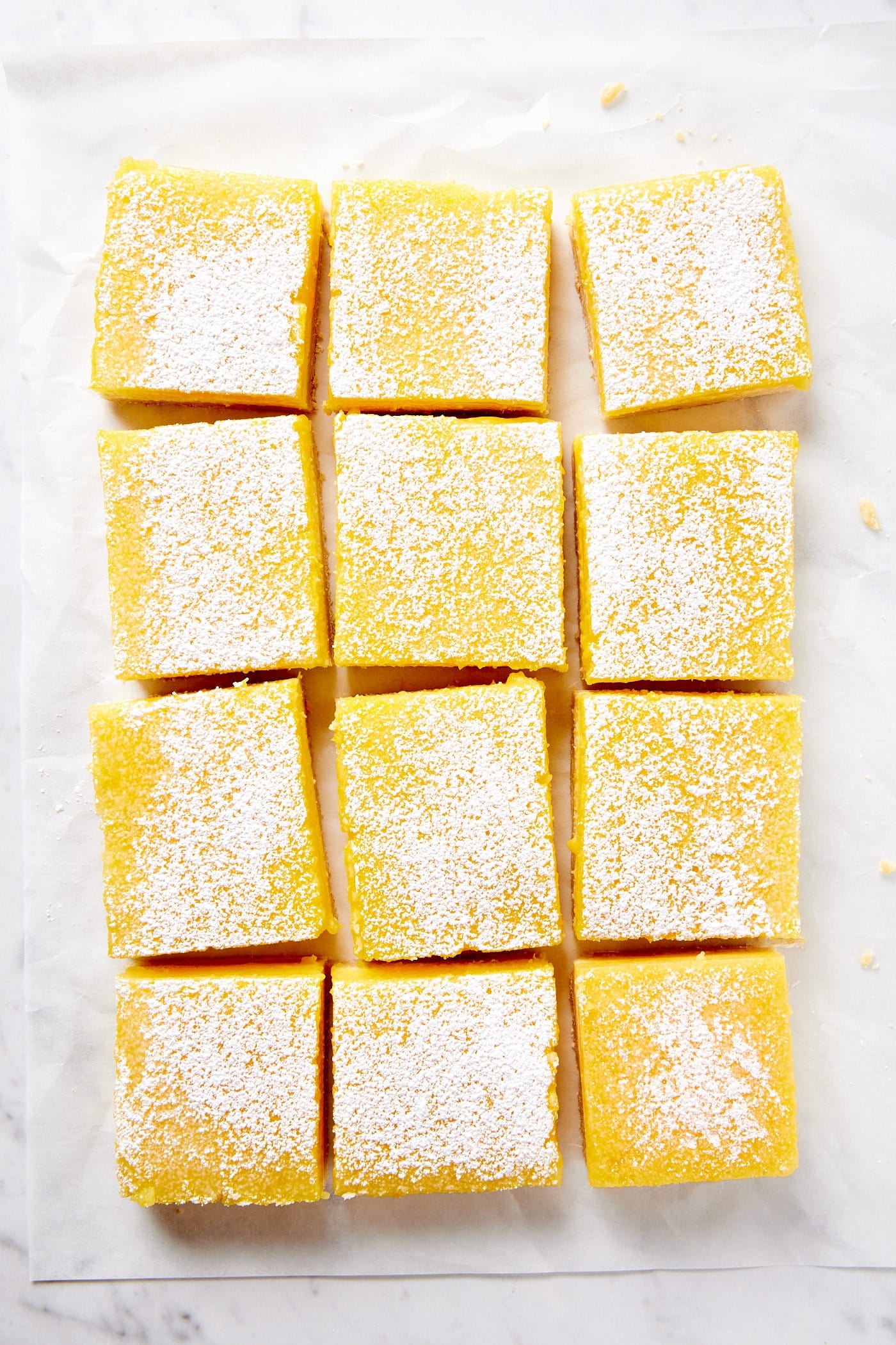 Squares of lemon cheesecake bars sitting on piece of parchment on marble surface.