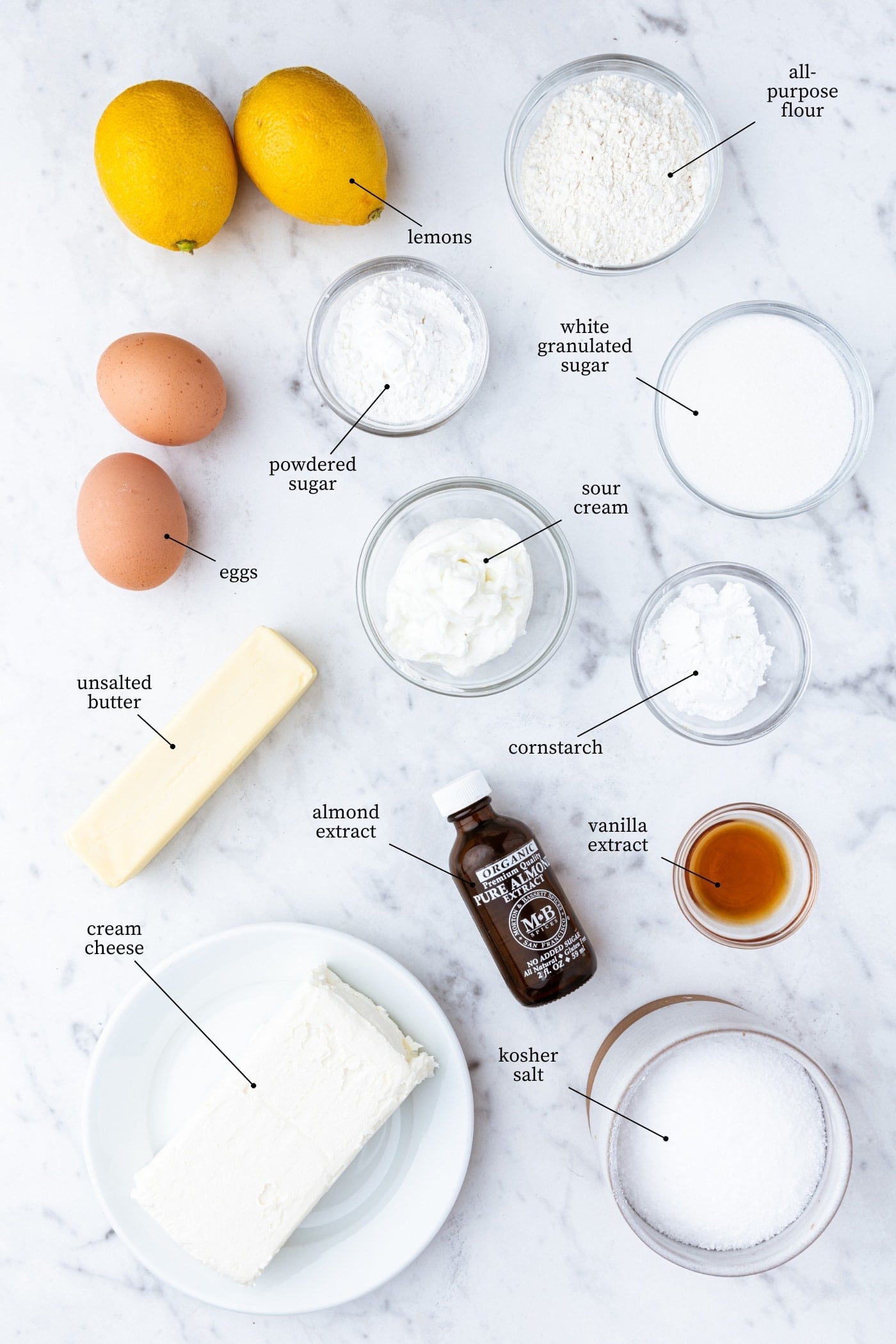White marble surface filled with ingredients needed to make lemon cheesecake bars including flour, sugar, eggs, lemons, spices, and more.