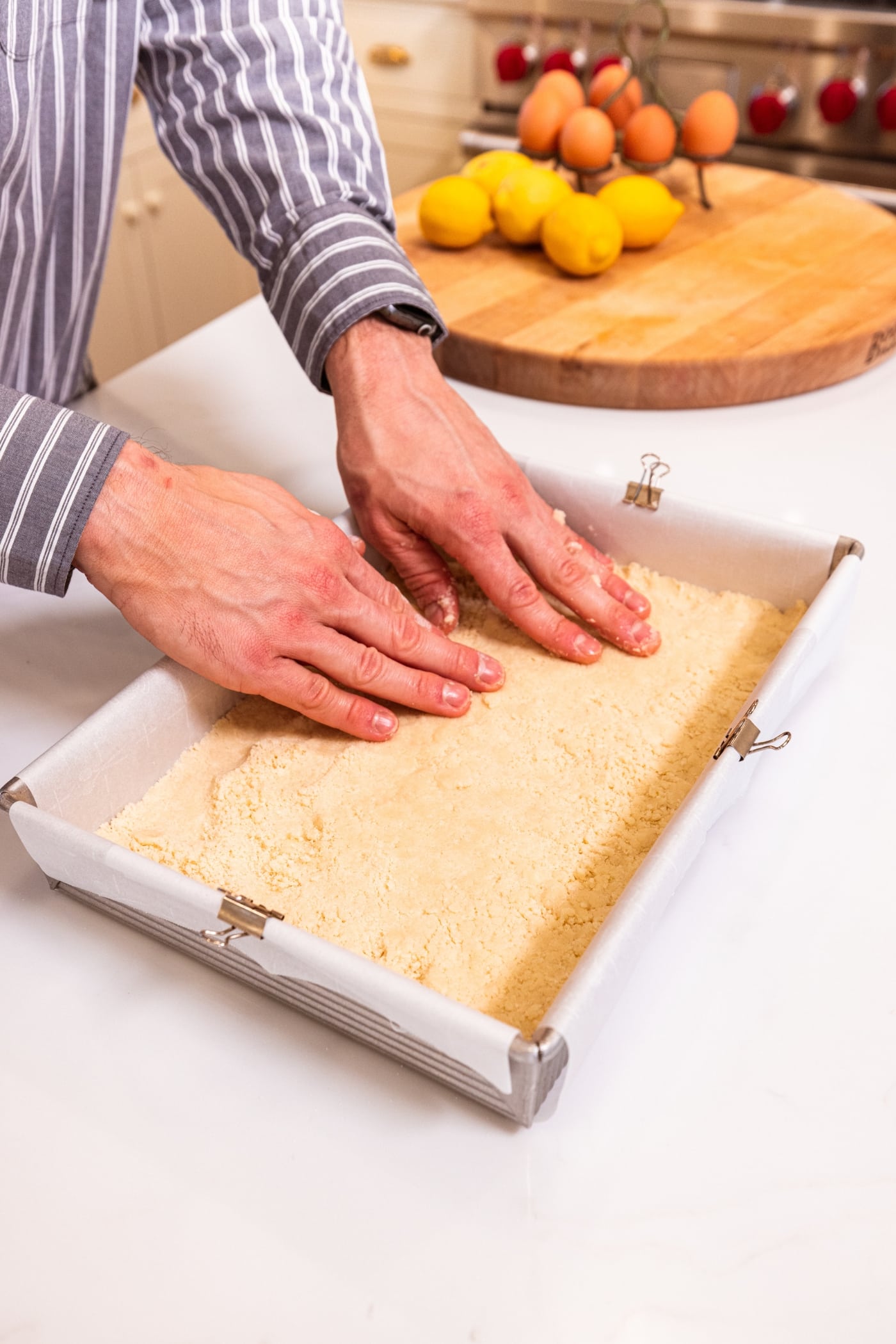 Two hands pressing shortbread crust into the bottom of a 9x13 baking dish with parchment and clips.