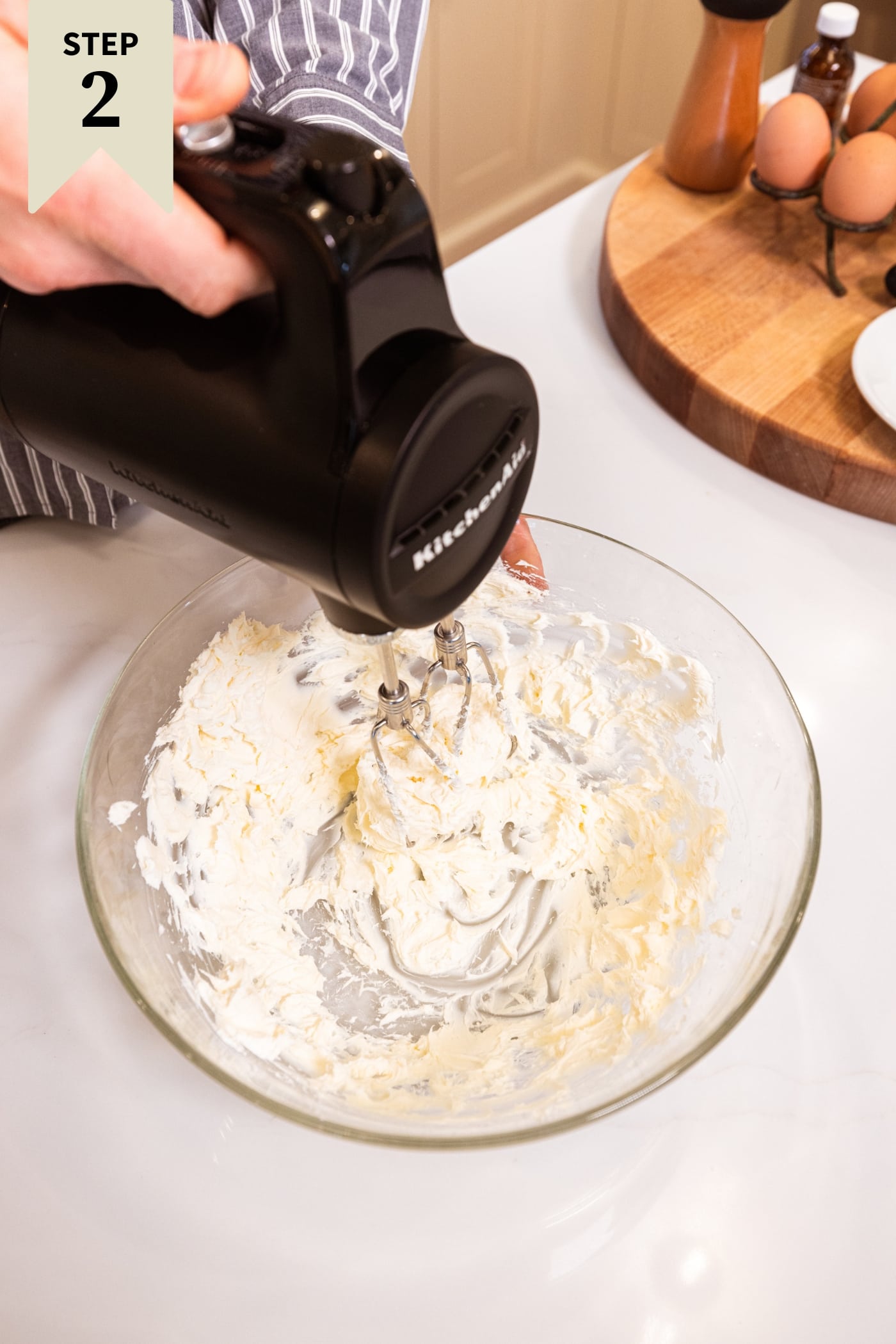 Handheld mixer mixing together softened cream cheese in a large glass bowl.