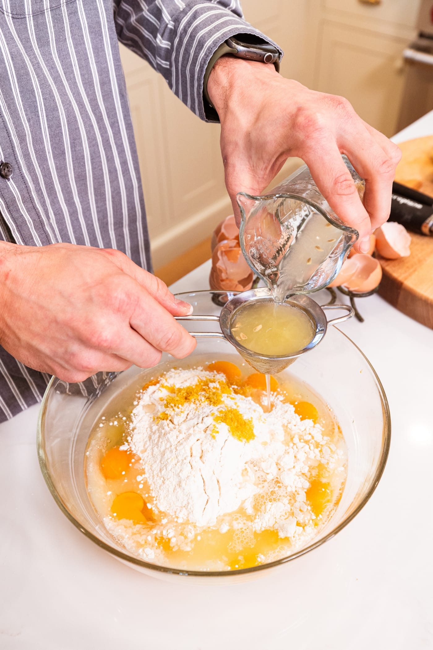 Hand holding strainer straining lemon juice into a glass bowl with ingredients for lemon juice filling.