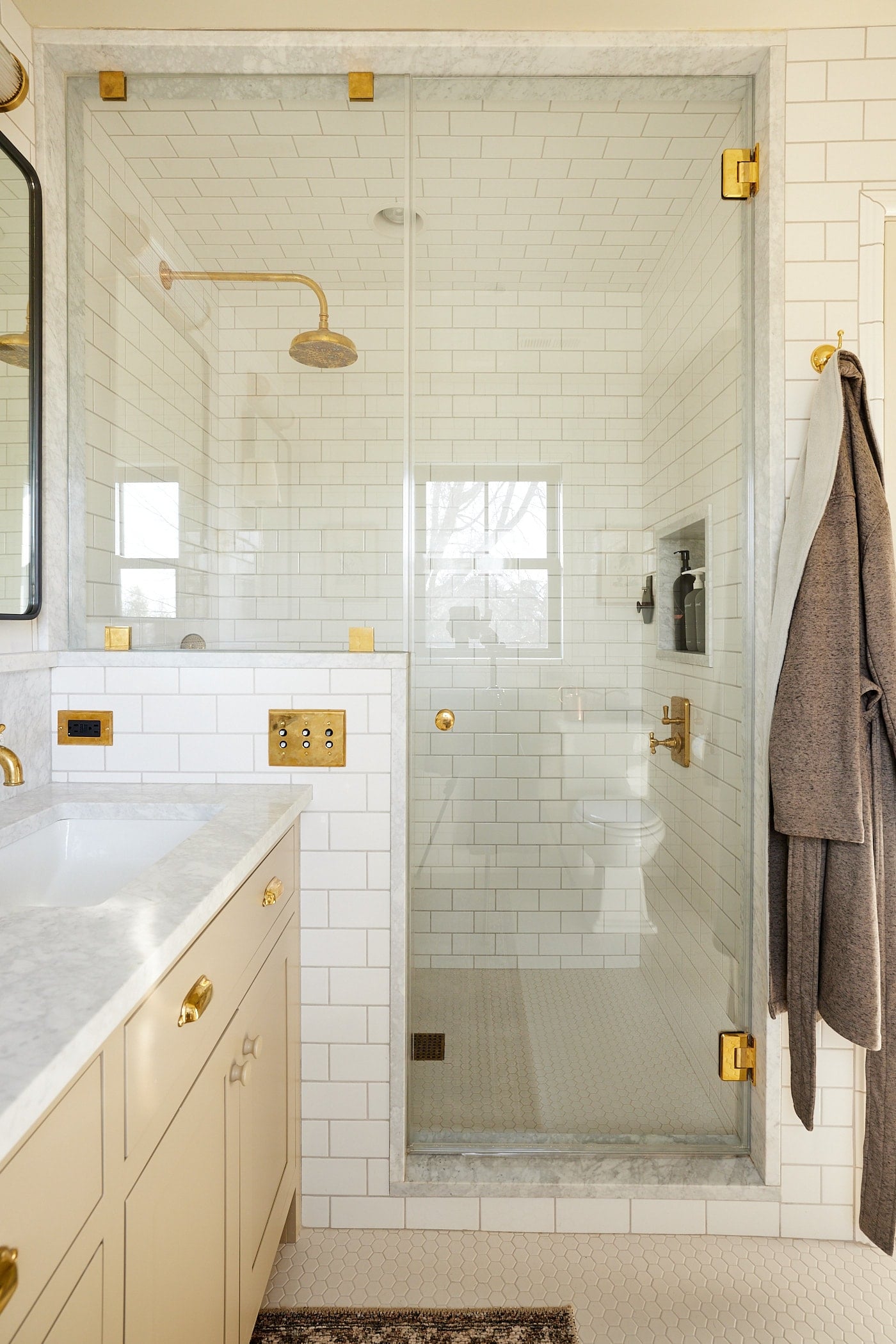 Glass and tile shower with brass fixtures with door right beside a khaki colored cabinet with mirror and a robe hanging to the side.