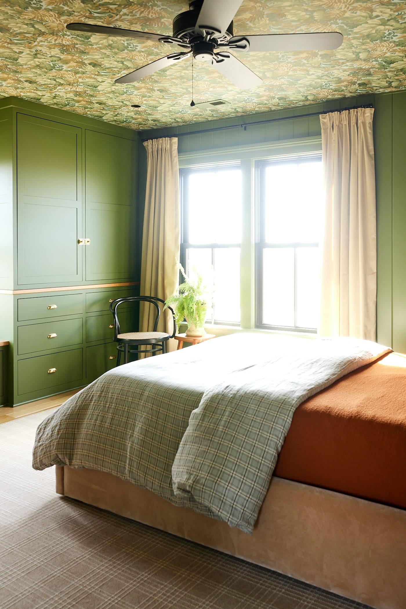 Green colored bedroom with large windows and built-in cabinets to the side with queen-sized bed with orange comforter and wallpaper on the ceiling.