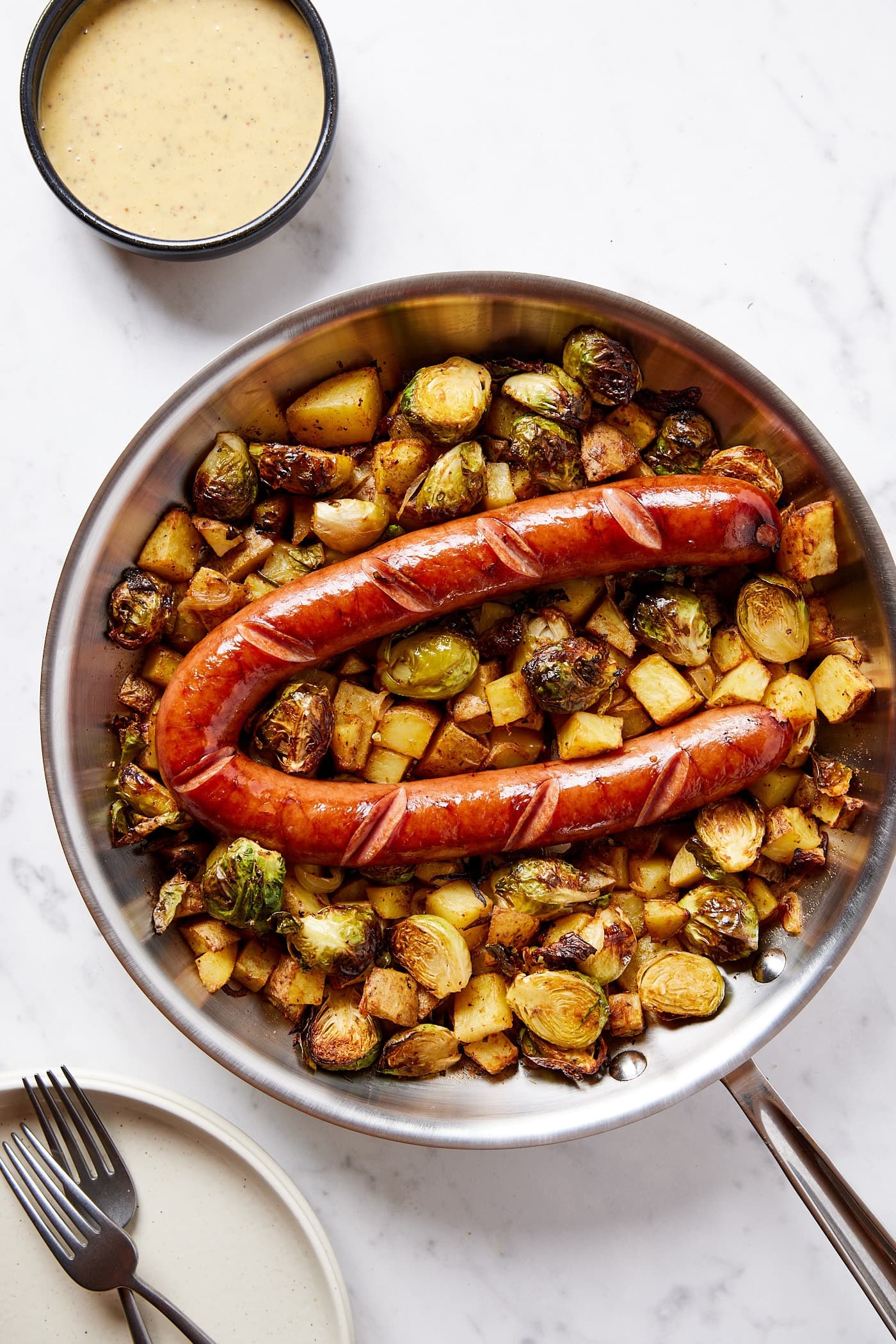 Metal skillet on white marble surface with mustard sauce to the side with more plates and forks.