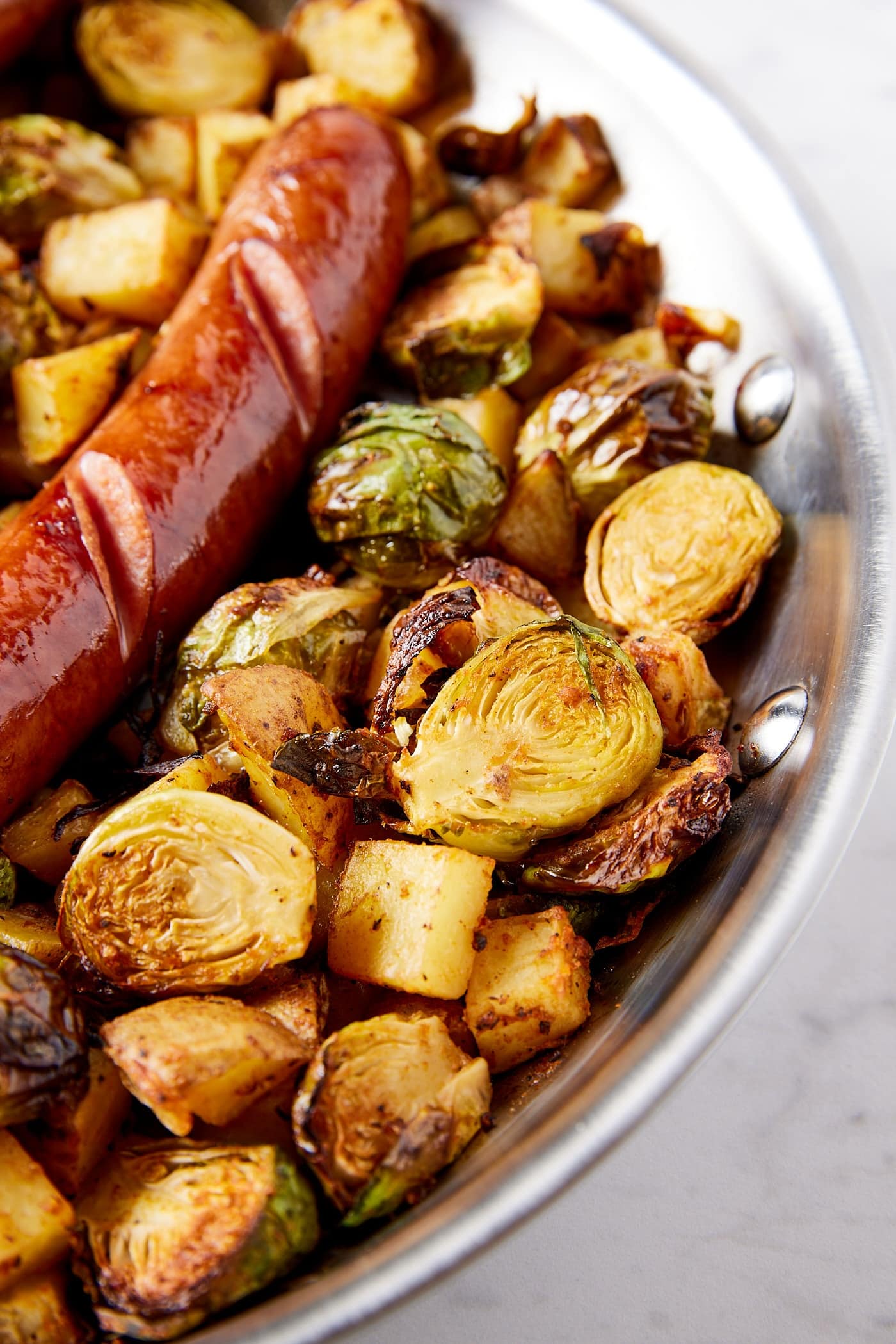 Metal skillet on marble surface filled with cooked Brussels sprouts and diced potatoes with kielbasa nestled on top.