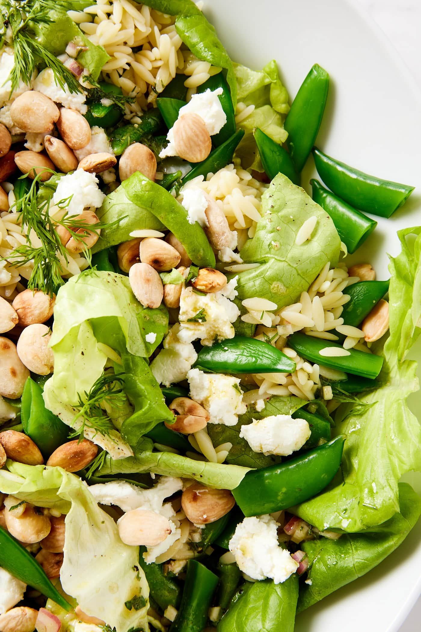 White salad bowl filled with spring orzo salad with toasted Marcona almonds, goat cheese, and blanched snap peas.
