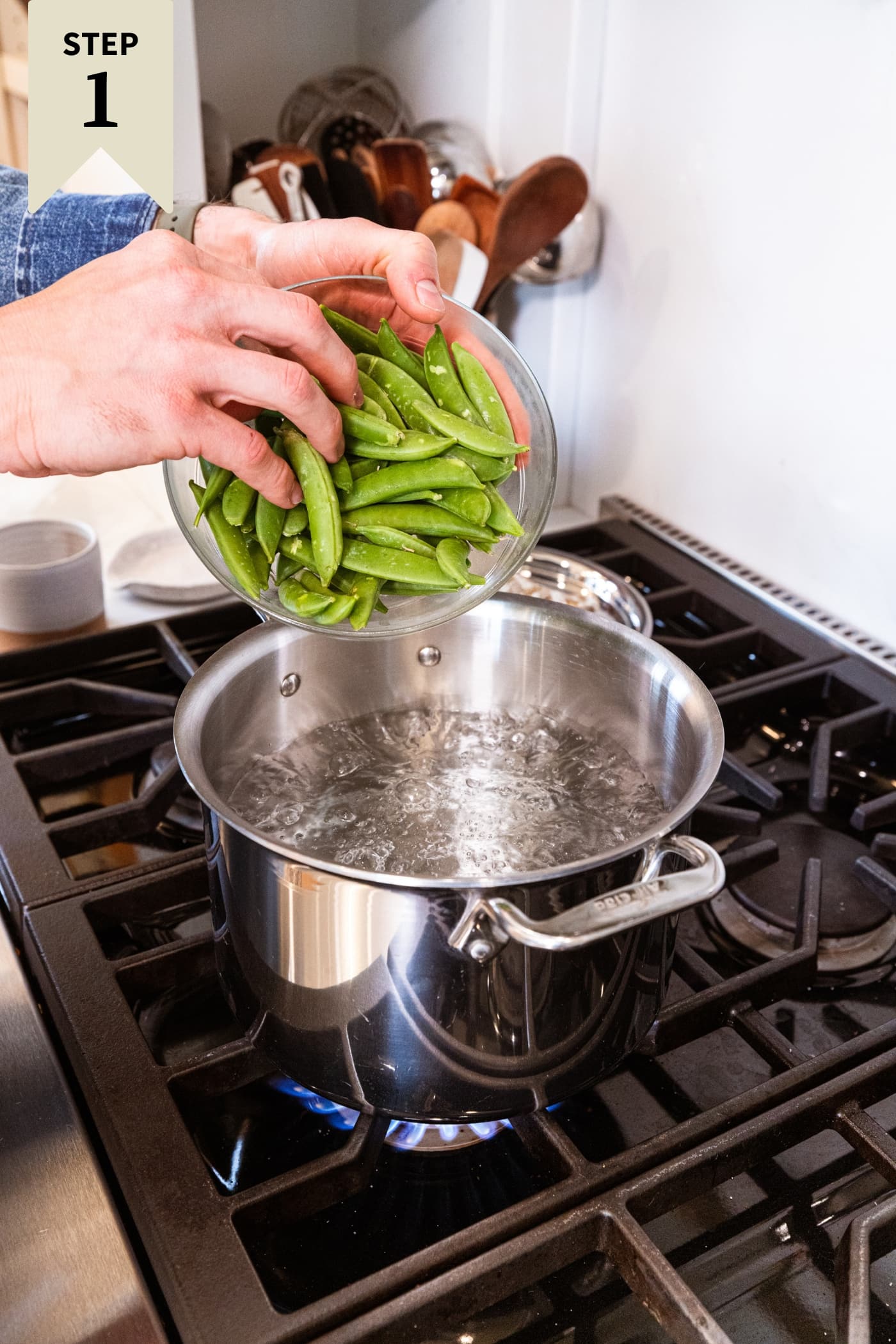 Hand holding glass bowl filled with snap peas about to pour into a large pot filled with boiling water to blanch them.