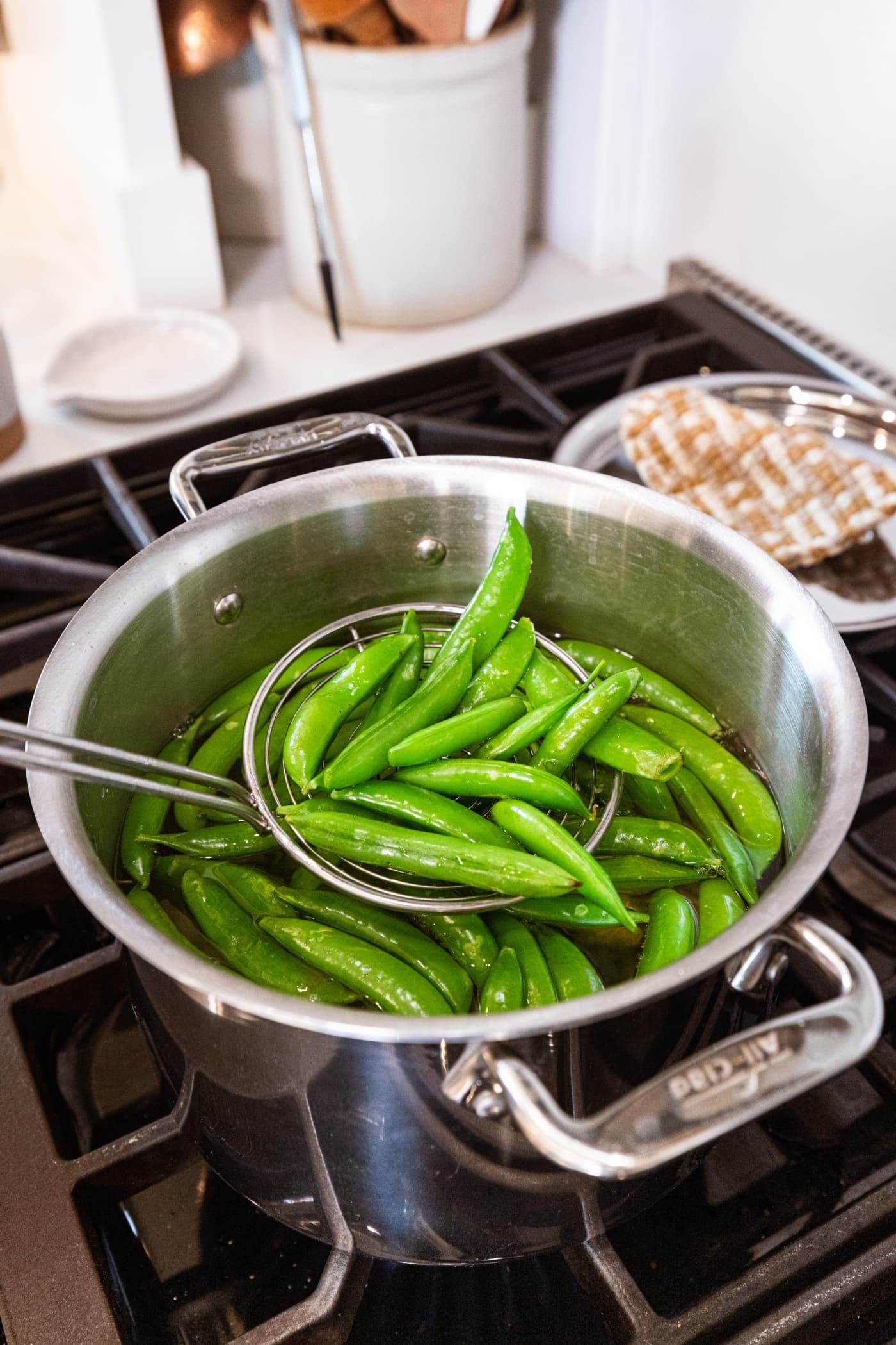 Ladle scooping up blanched snap peas from boiling water in a large pot on the stove.
