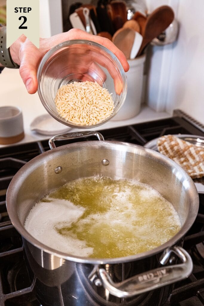 Hand holding glass bowl pouring raw orzo into a large pot filled with boiling water already with orzo inside.