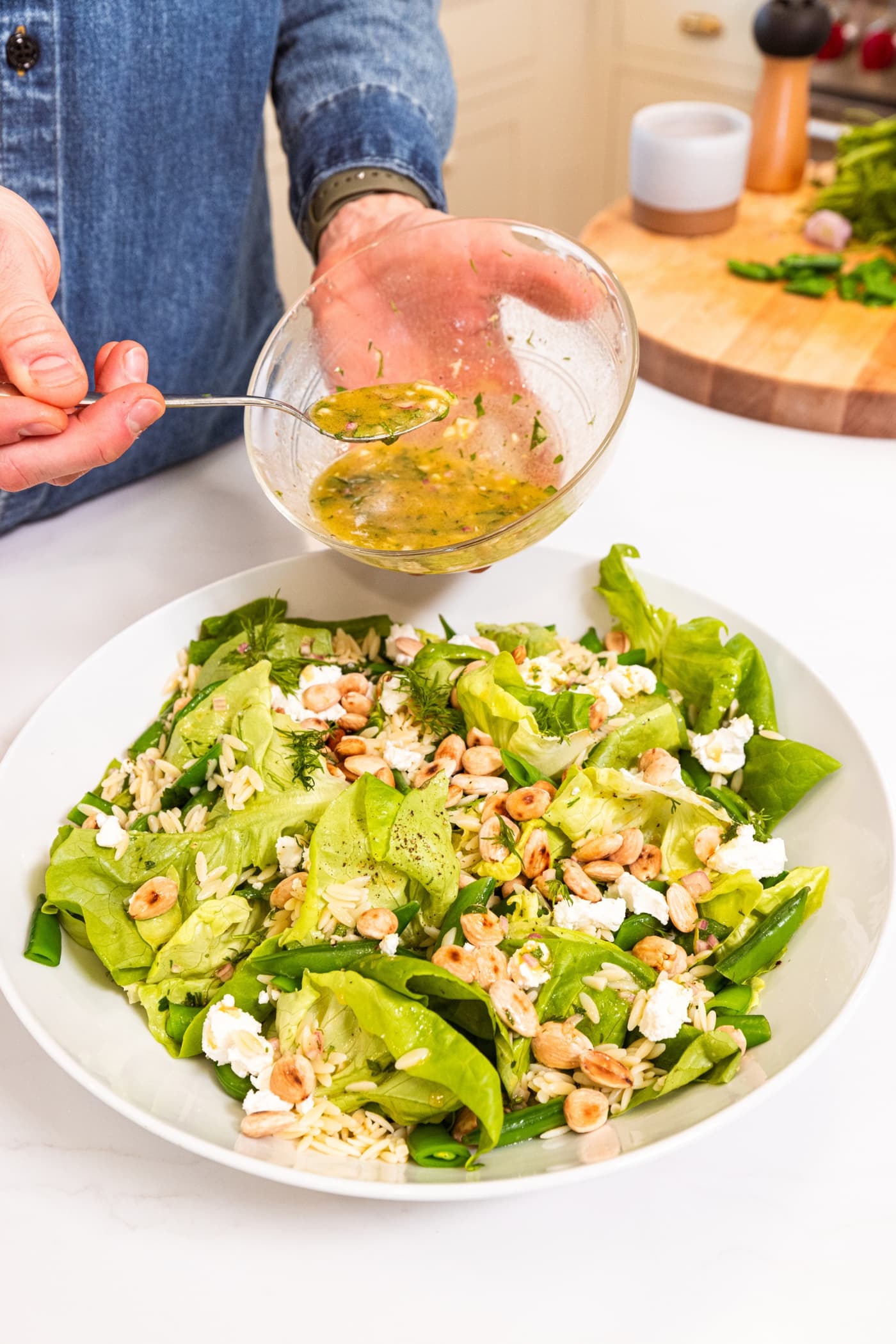 Hand holding glass bowl drizzling salad dressing on top of spring orzo salad with goat cheese and toasted Marcona almonds.