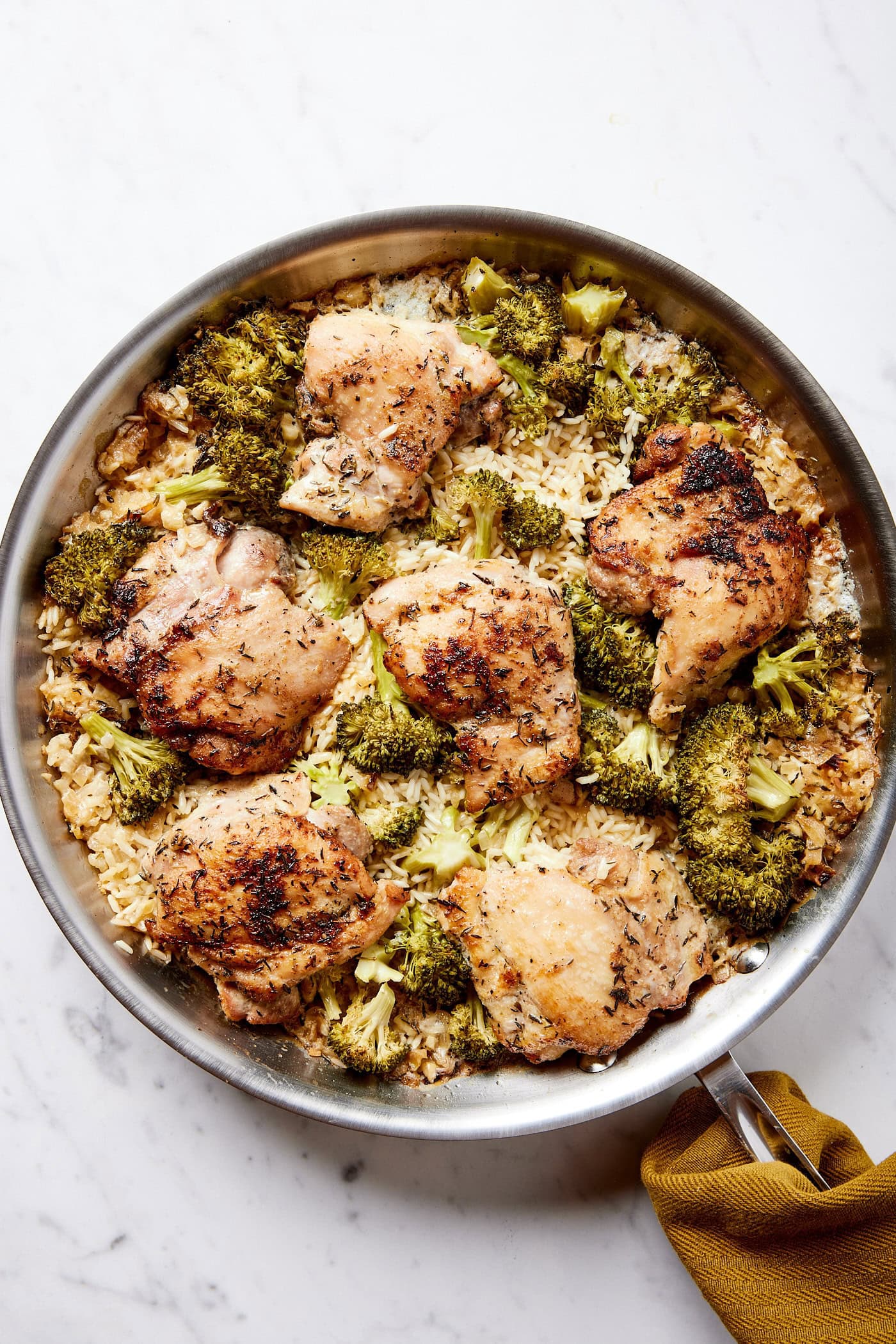 Metal skillet on white marble surface filled with cooked chicken, broccoli, and rice casserole with browned chicken texture.