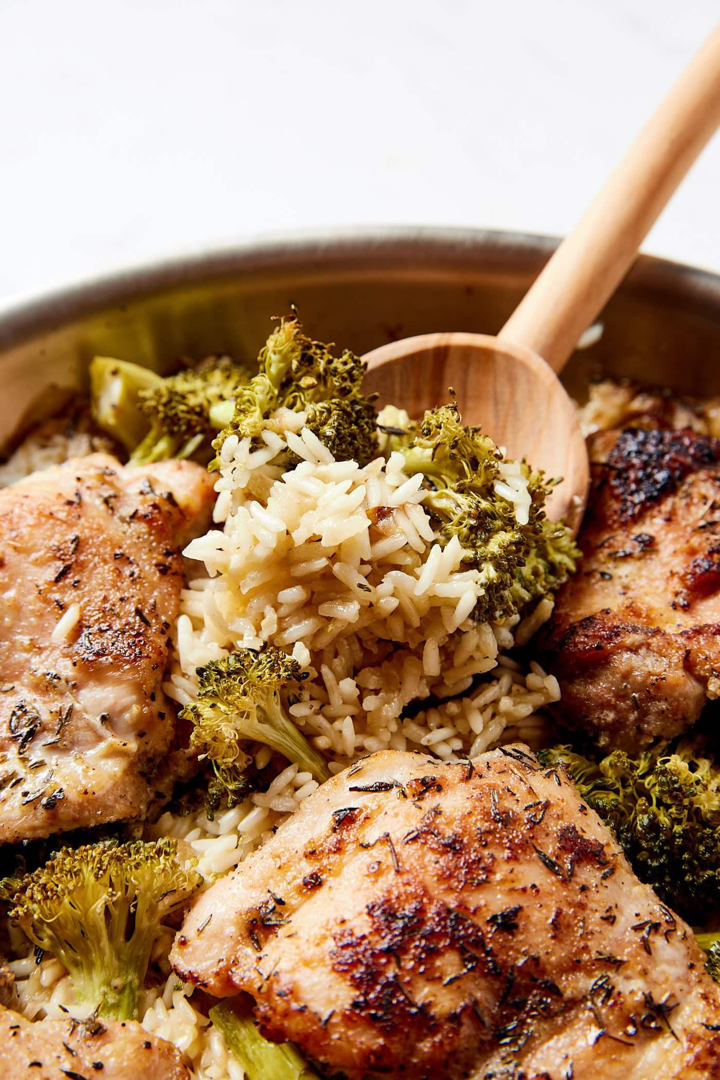 Wooden spoon scooping up a serving of rice with broccoli from a metal skillet filled with chicken thighs.
