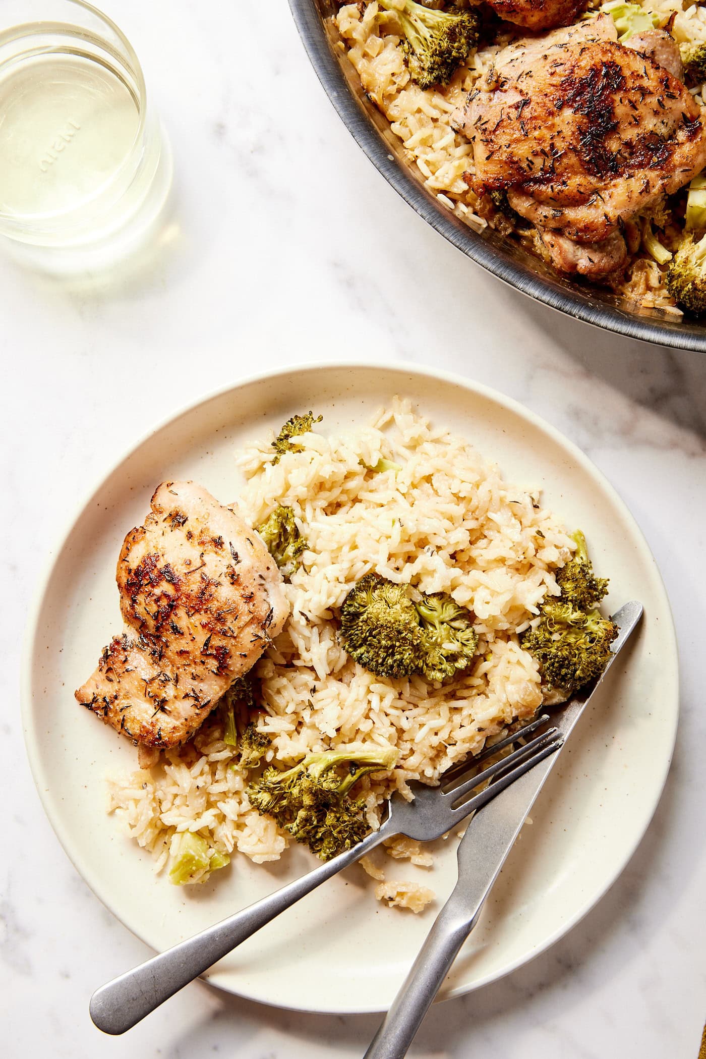 White marble surface with plate with serving of chicken and rice and broccoli with utensils resting on the plate.