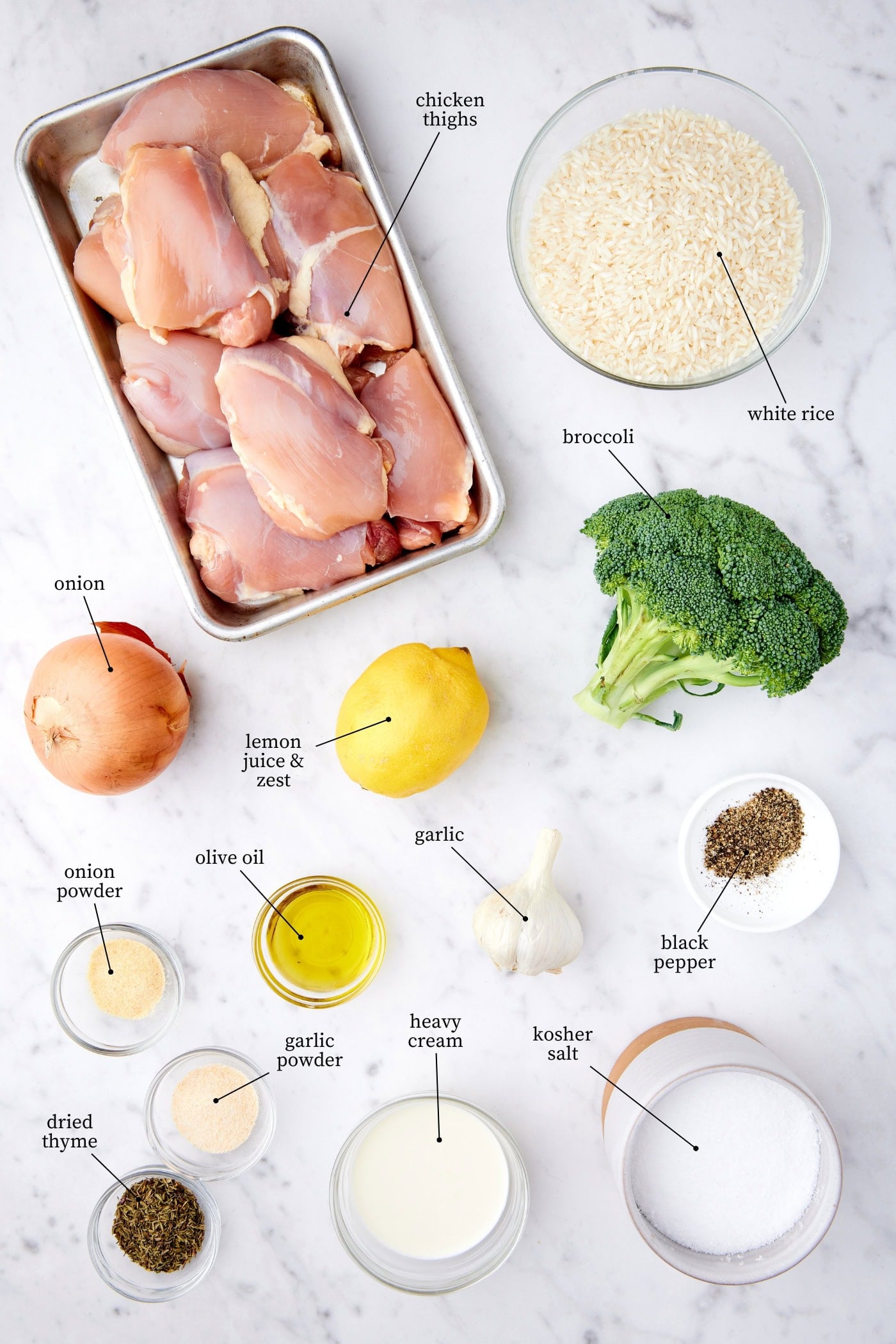 White marble surface filled with ingredients needed to make chicken, broccoli, rice casserole including raw chicken, broccoli, rice, onion, lemon, spices, and more.