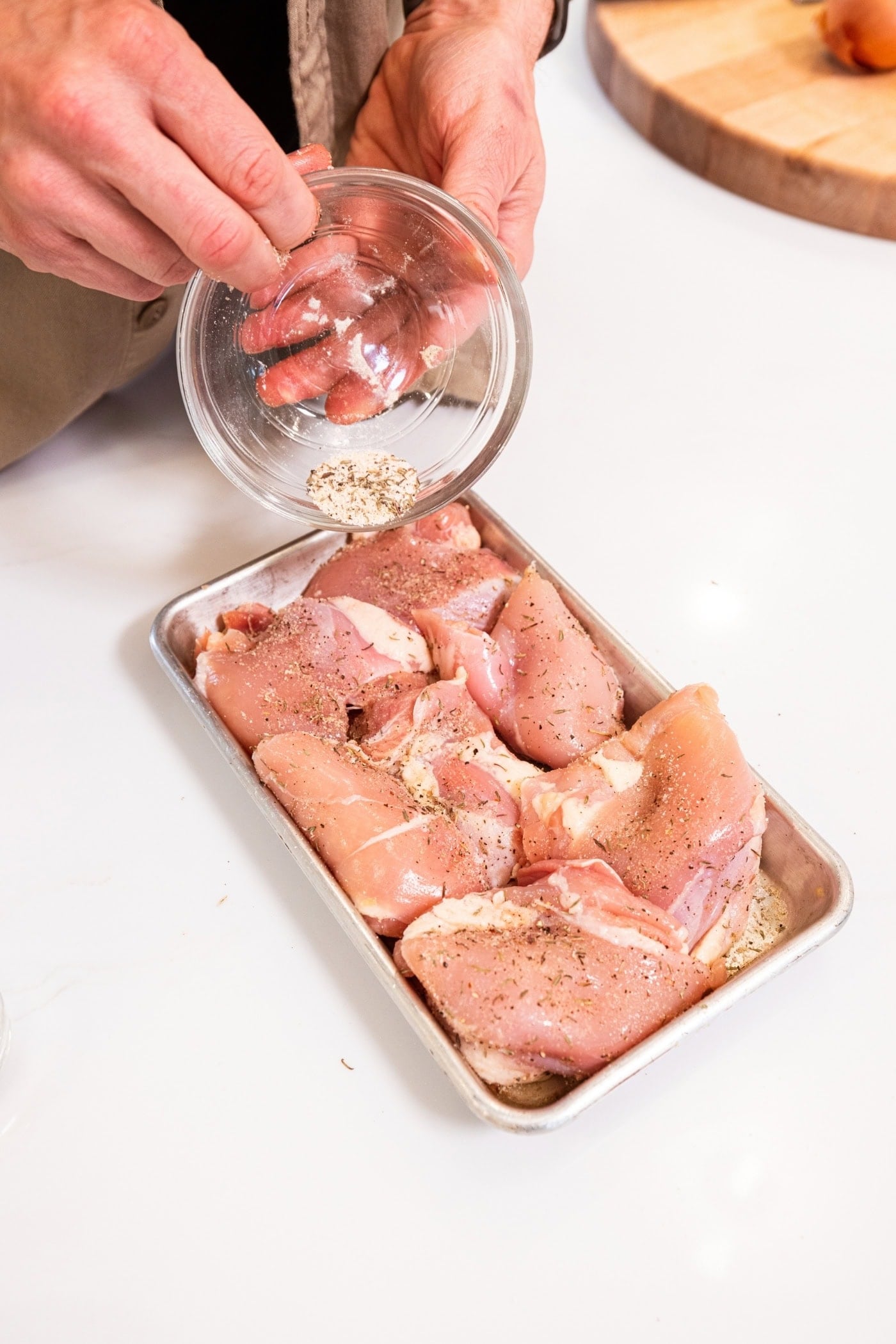 Hand pouring dry rub on top of raw chicken thighs on a small metal sheet before letting marinate.