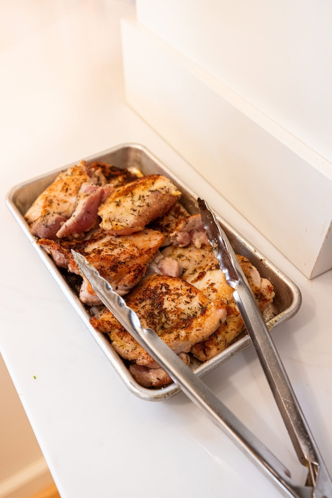 Chicken thighs after being browned on one side sitting on a metal pan with tongs.