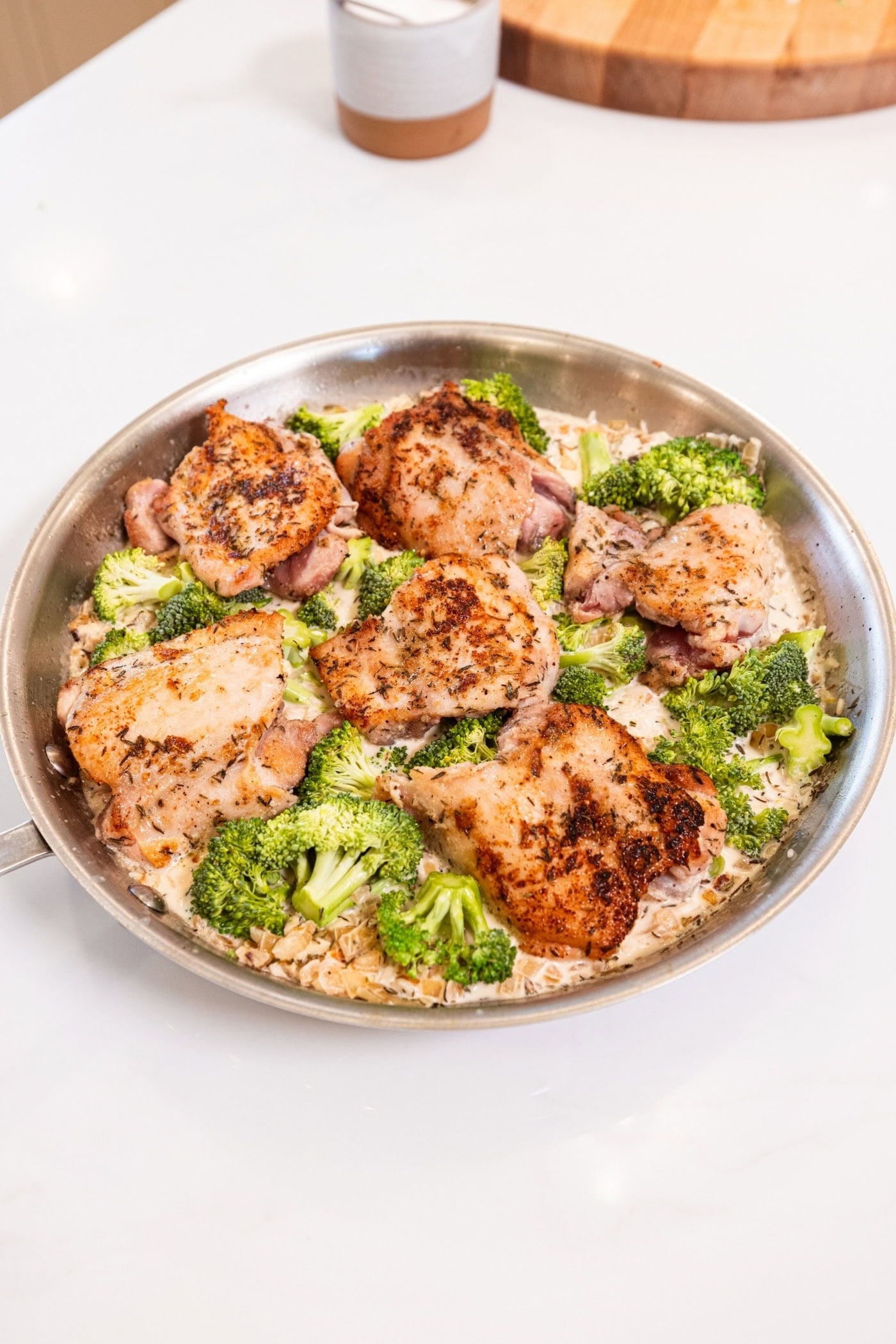 Metal skillet on a white surface filled with partially cooked chicken thighs and rice and broccoli.