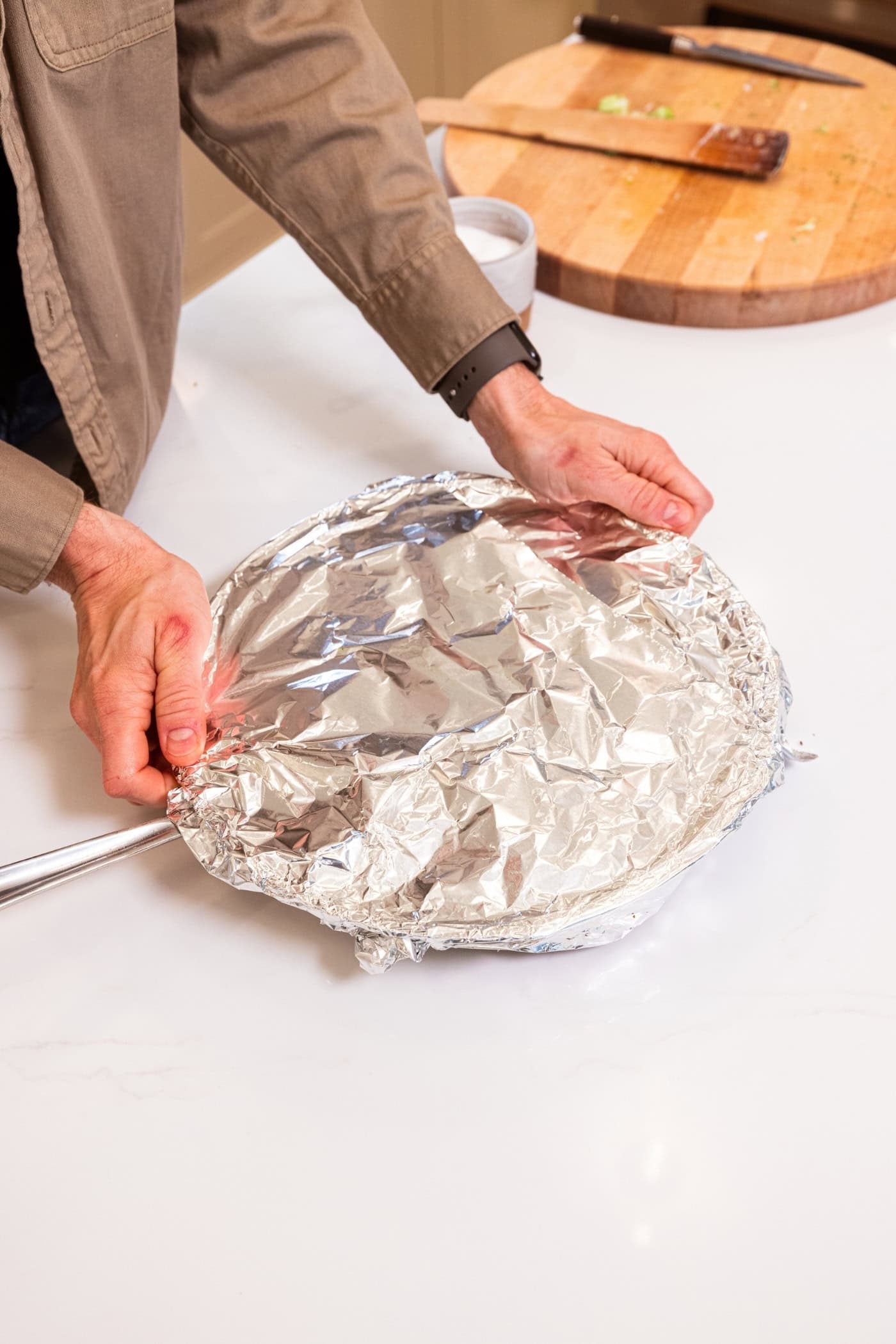 Two hands placing aluminum foil over the top of a skillet with chicken.