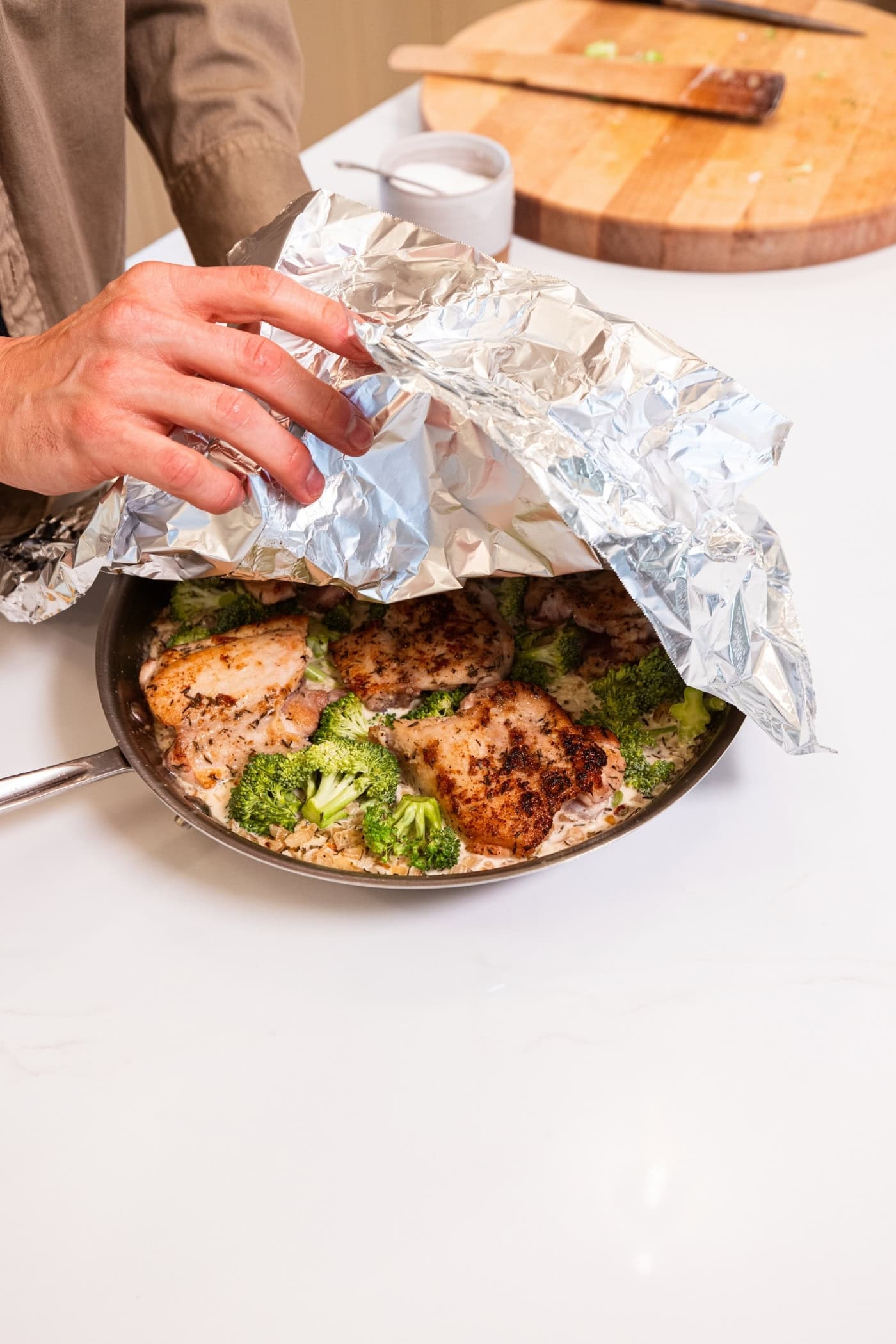 Hand pulling back cover on skillet filled with cooking chicken, broccoli, and rice.