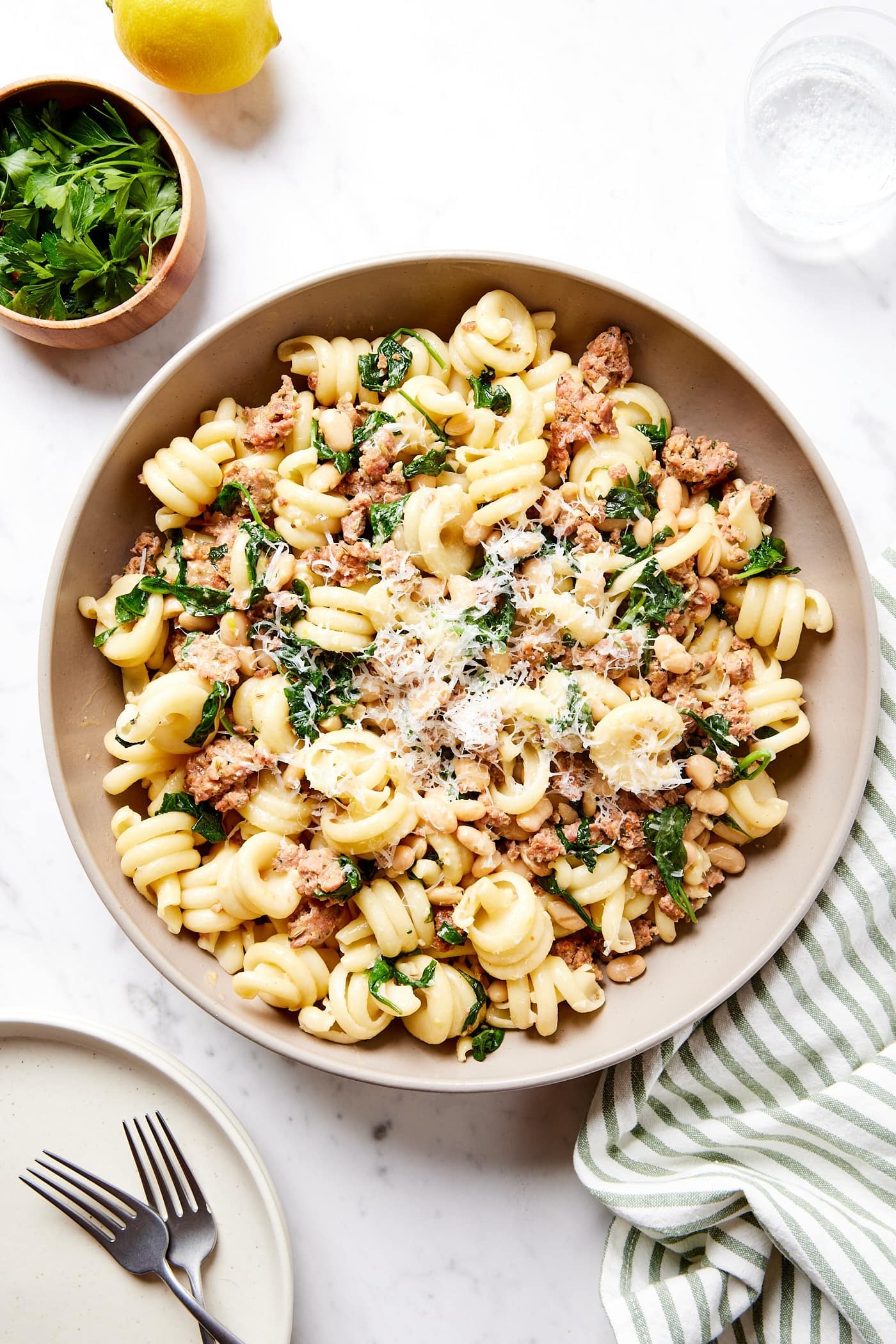 Marble surface with brown bowl filled with weeknight sausage pasta with herbs and lemon to the side along with plates for serving.