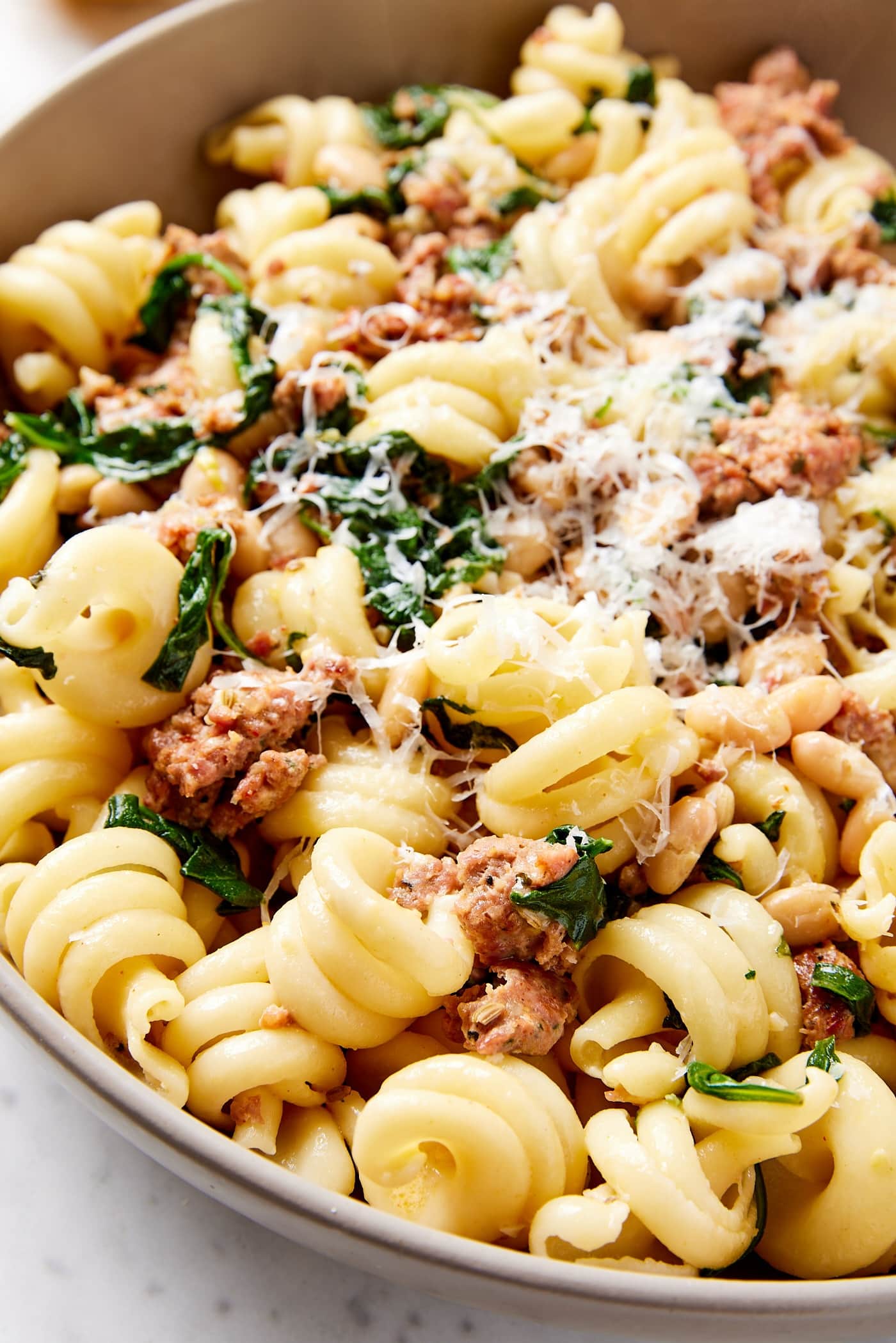 Brown bowl on marble surface packed with cooked twirls of pasta topped with Parmesan cheese, spinach, and cooked sausage for a weeknight meal.