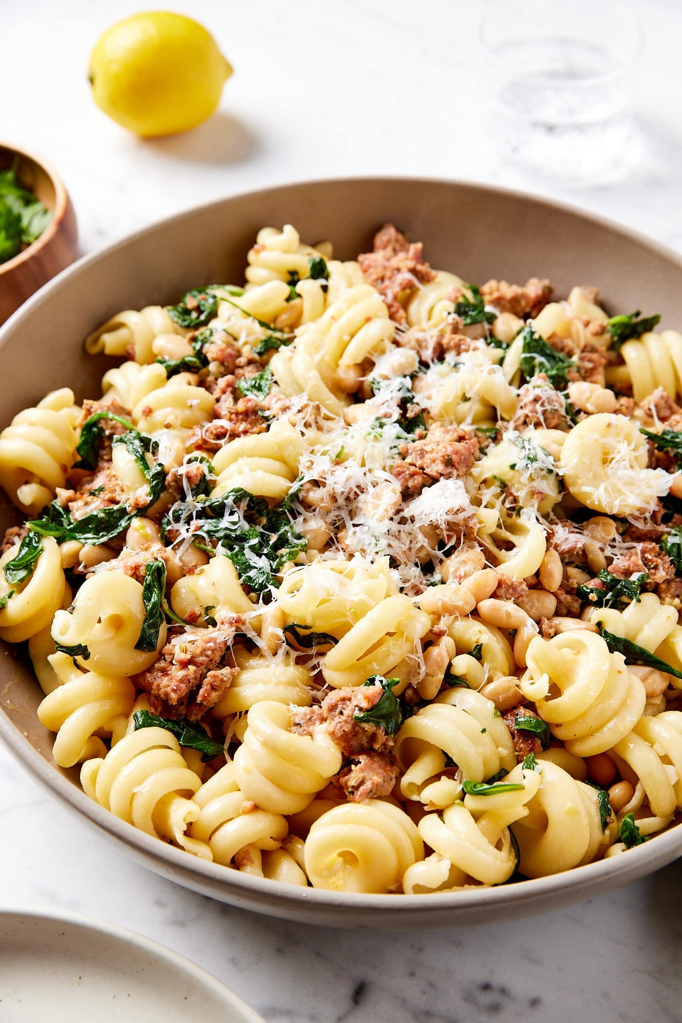 Brown bowl filled with cooked mixture of pasta with spinach and sausage and white beans with lemon in the background.