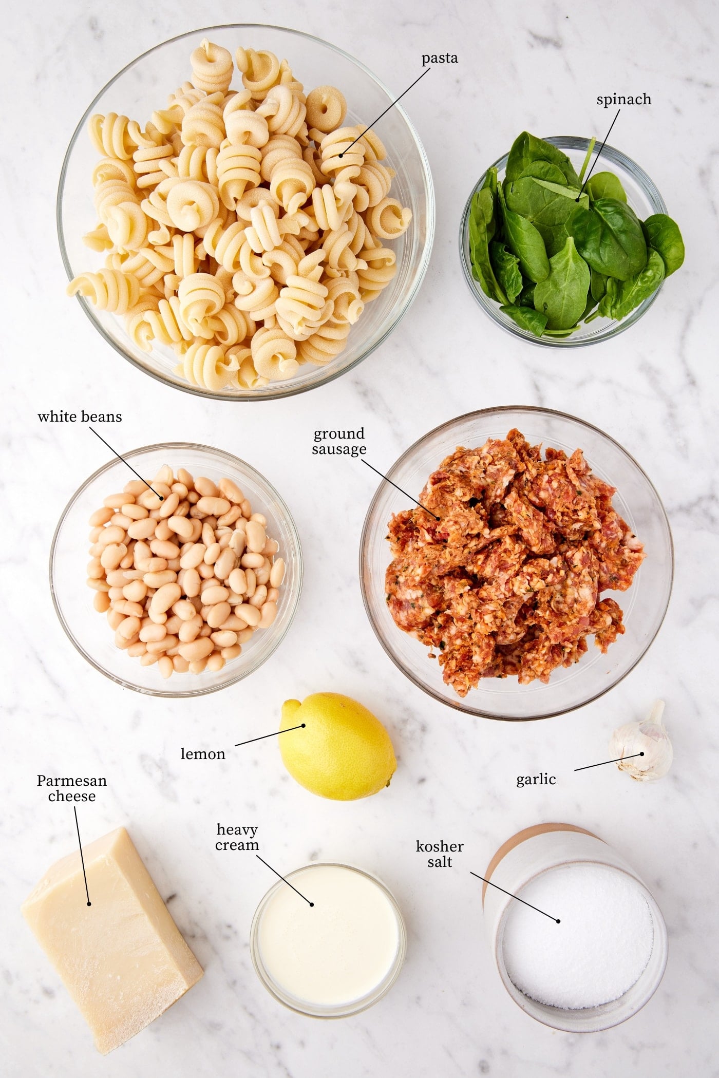 Marble surface filled with all ingredients needed to make spinach & sausage pasta including pasta, spinach, sausage, lemon, and spices.