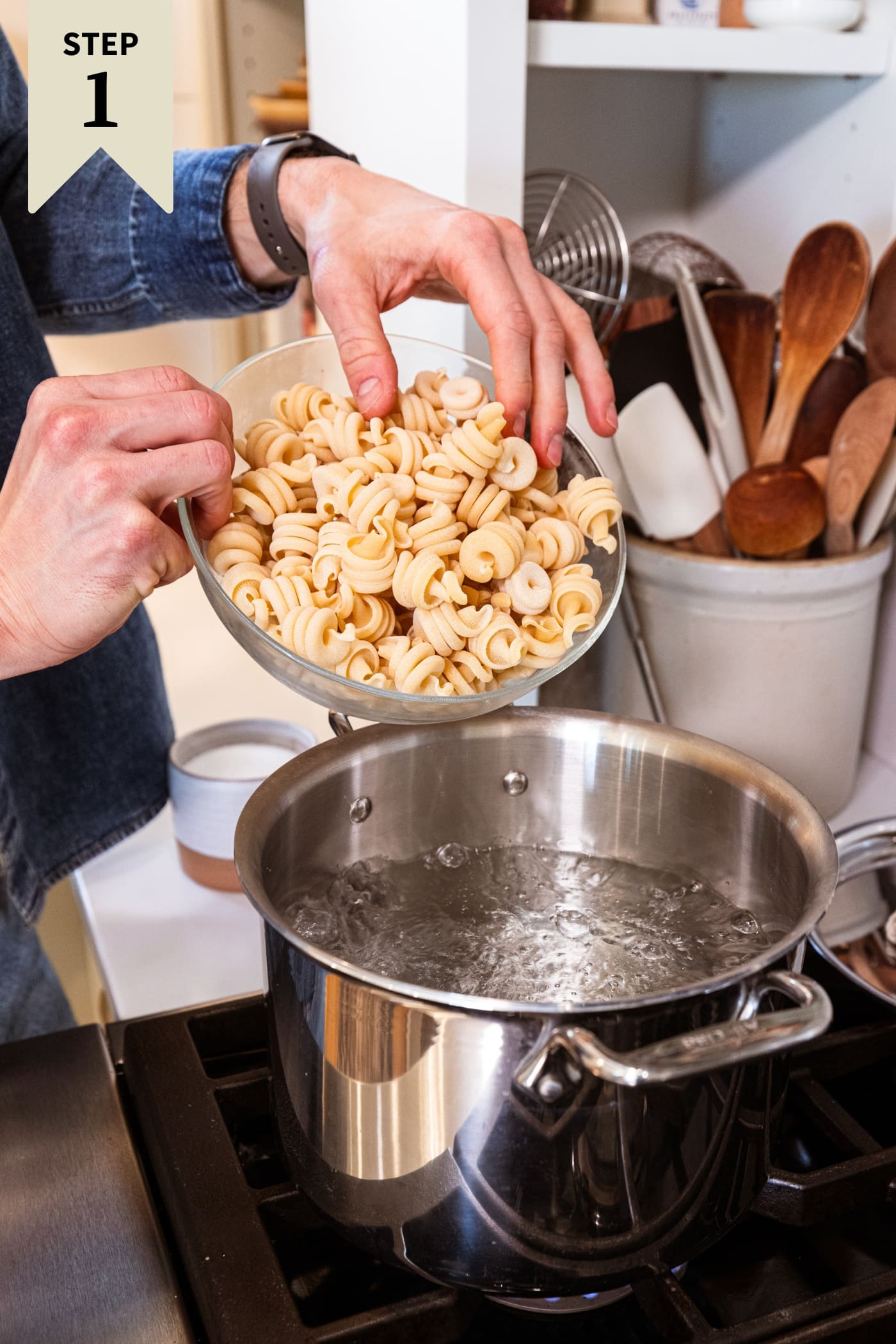 Hand about to pour uncooked twirled pasta into a large pot on the stovetop filled with boiling water to cook.