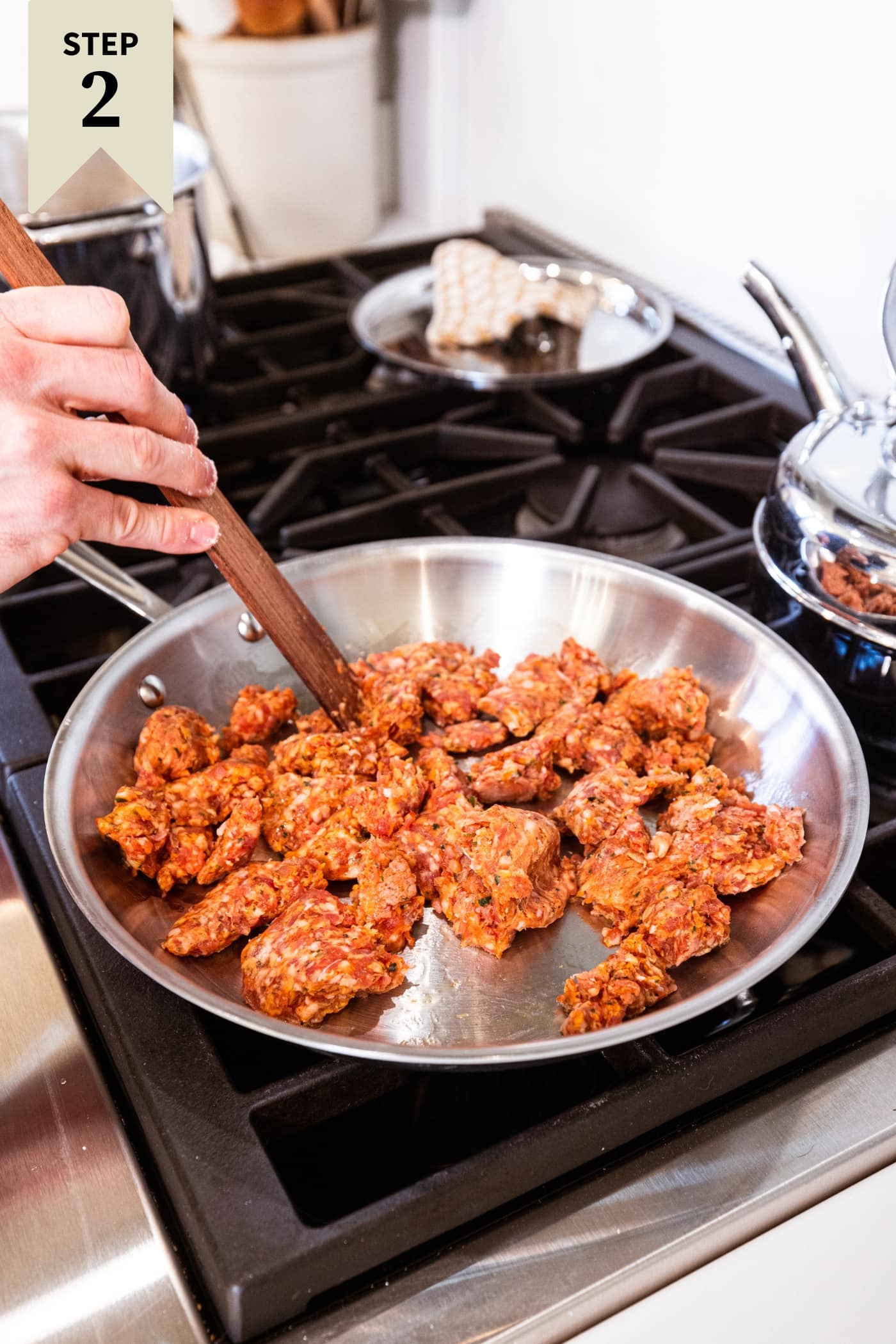 Metal skillet with uncooked sausage being mixed together with wooden spatula on stovetop.