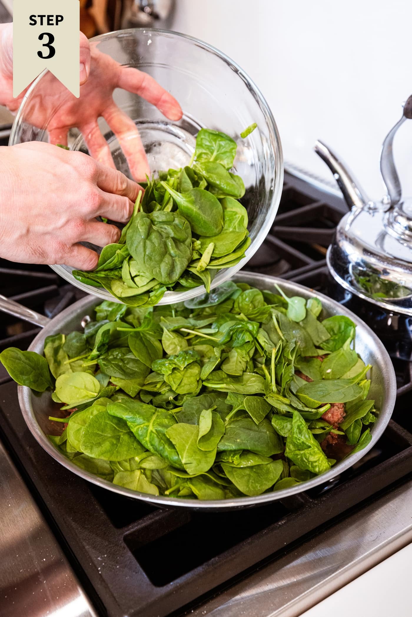 Hand pouring spinach from glass bowl into a skillet with meat to let cook down and decrease in volume.