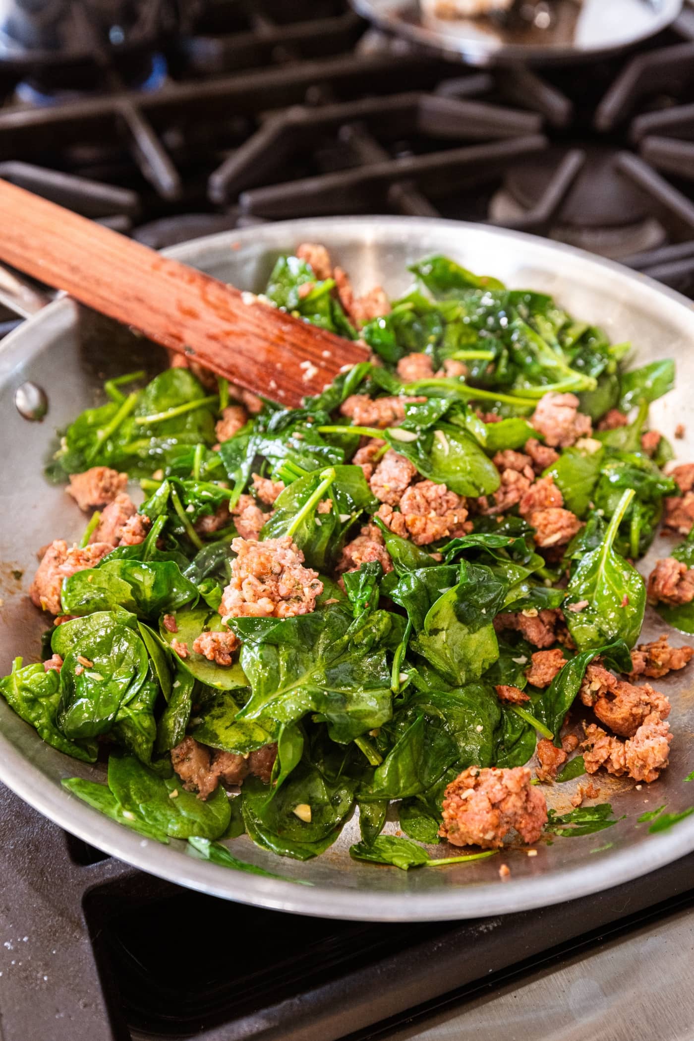 Mixture of cooking spinach and sausage in a metal skillet on the stovetop.