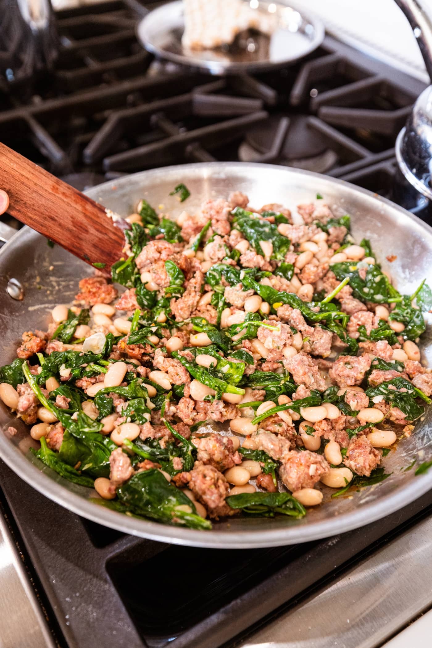 Mixture of cooked sausage with wilted spinach and white beans all in a metal skillet on the stovetop.