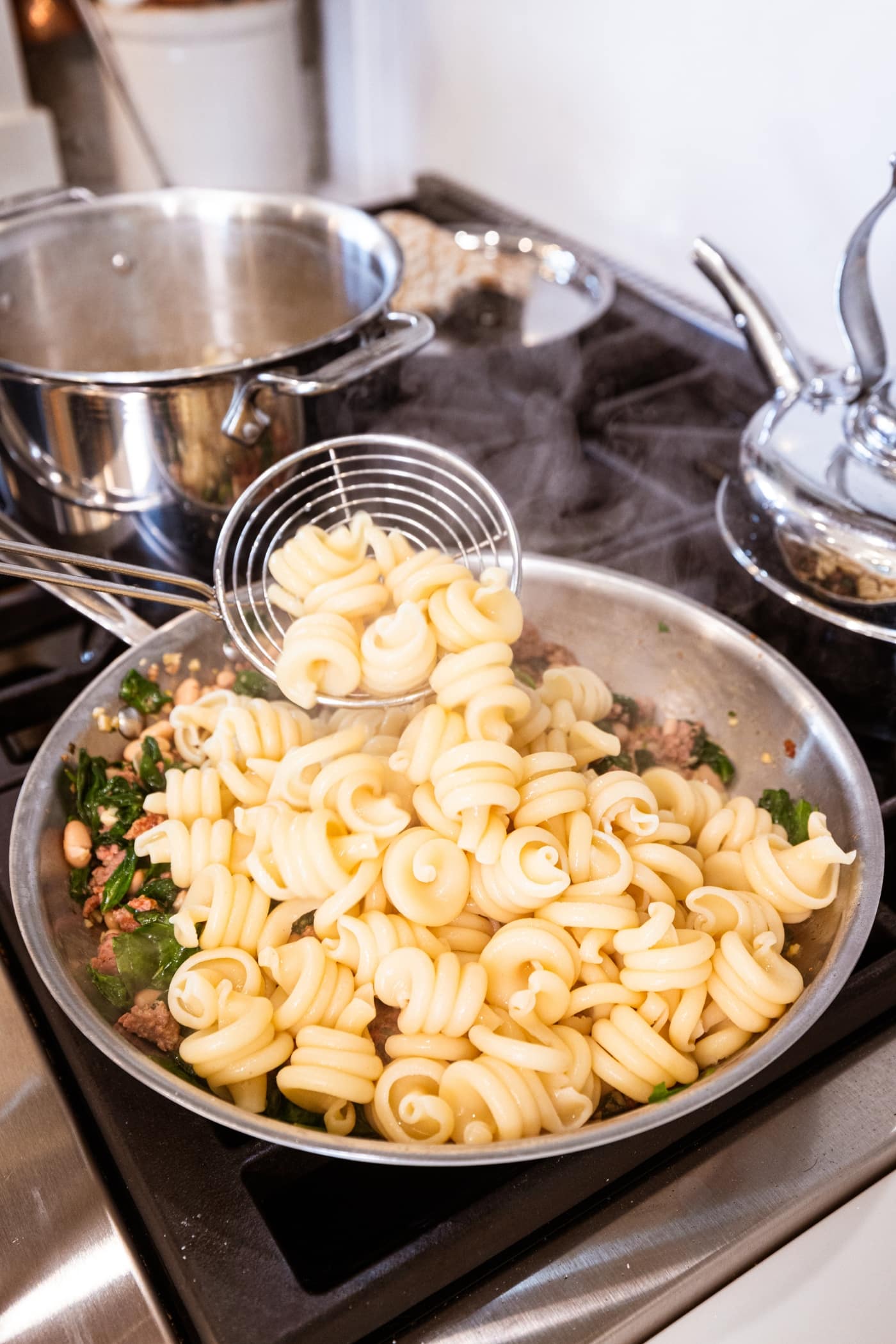Ladle scooping cooked pasta on top of sausage and spinach mixture in a metal skillet on the stovetop.