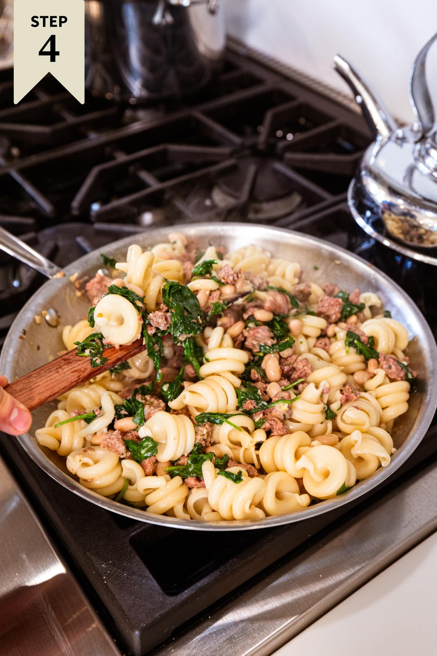 Metal skillet on the stovetop with cooked pasta and spinach and sausage after being mixed together and warmed slightly.