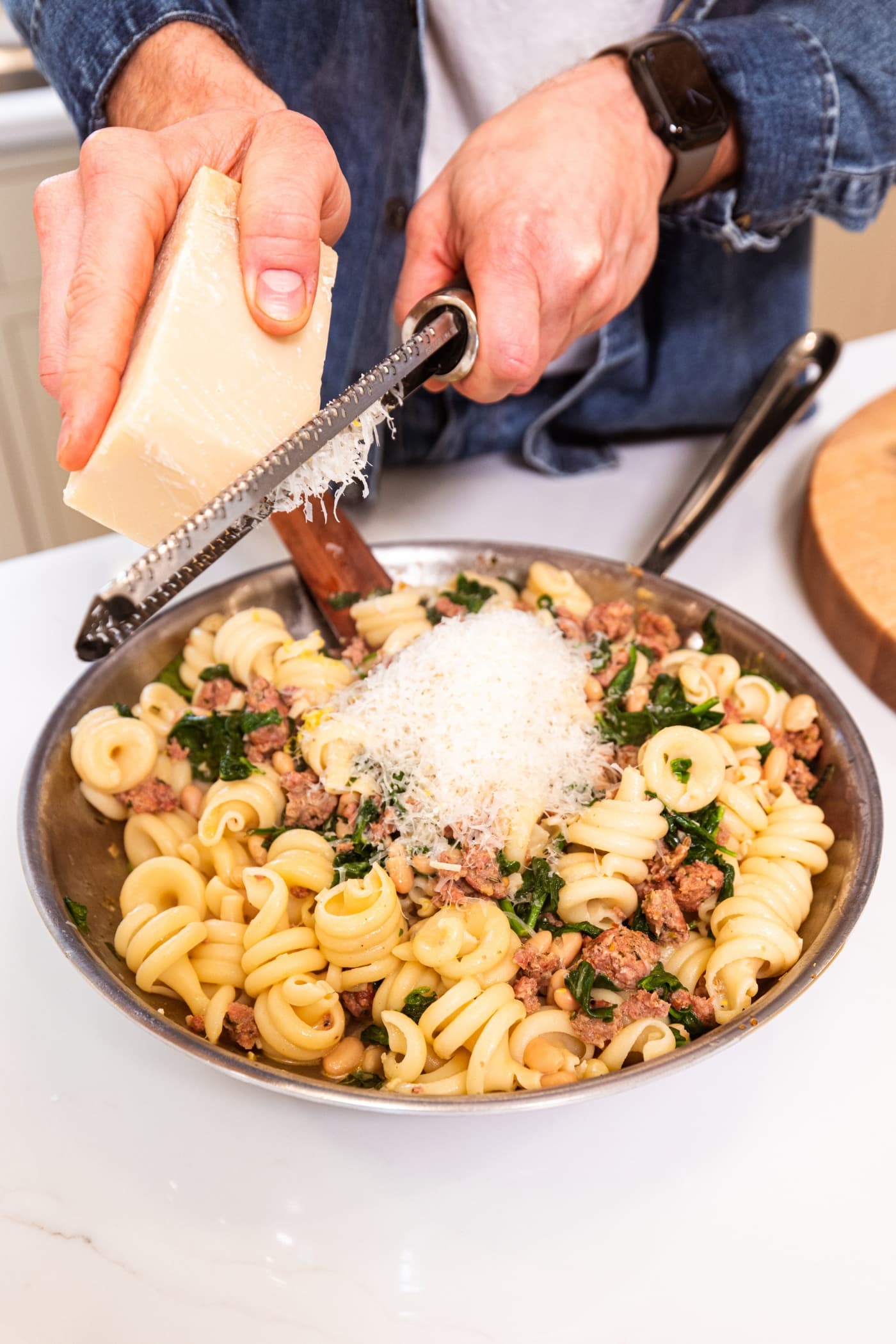 Hand using Microplane to grate Parmesan cheese on top of pasta and sausage mixture in metal skillet.